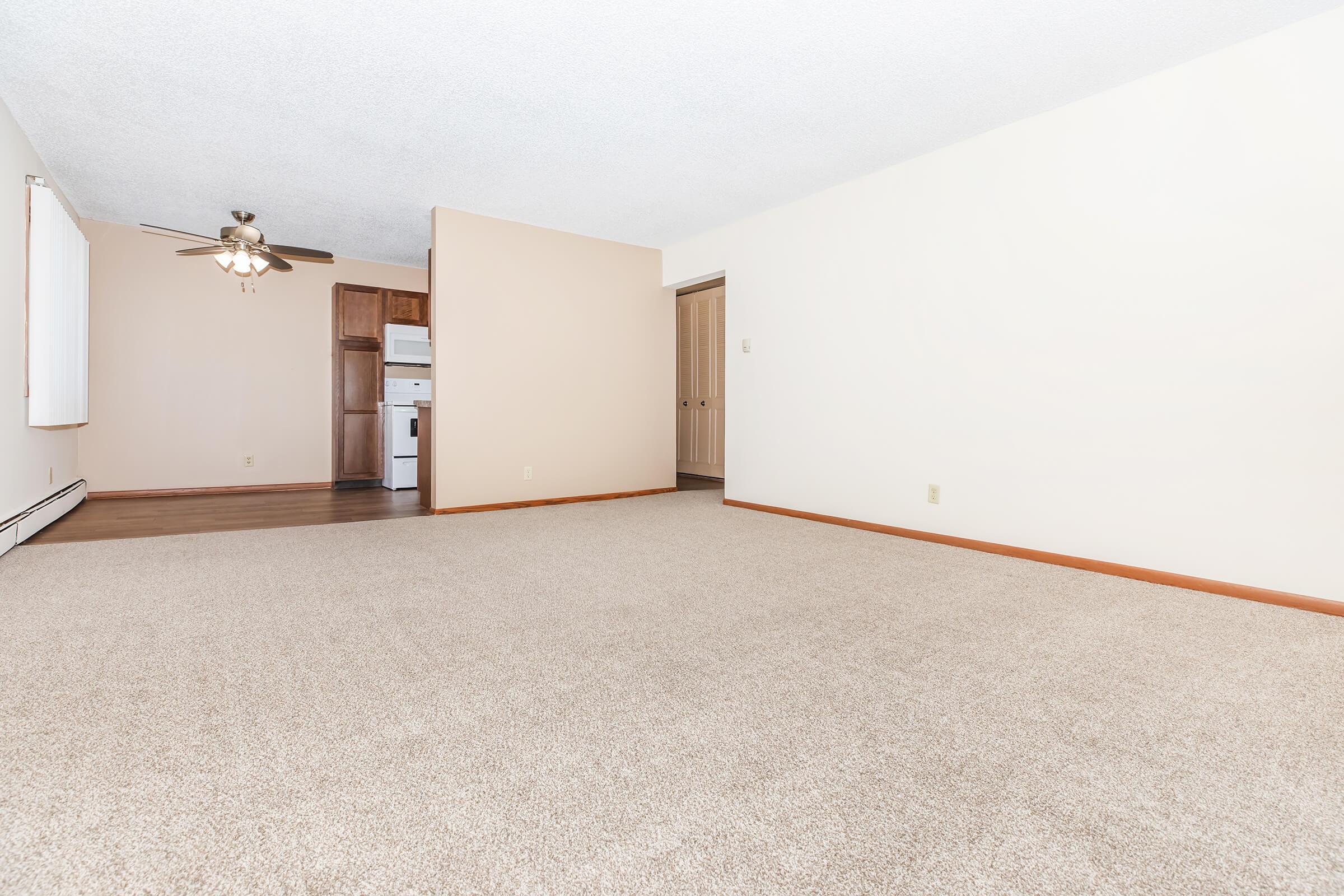 A spacious, empty living room with beige carpet and light-colored walls. A ceiling fan is visible, and there's a doorway leading to a kitchen area with white appliances. The room has ample natural light and a neutral color palette, creating a bright and airy atmosphere.