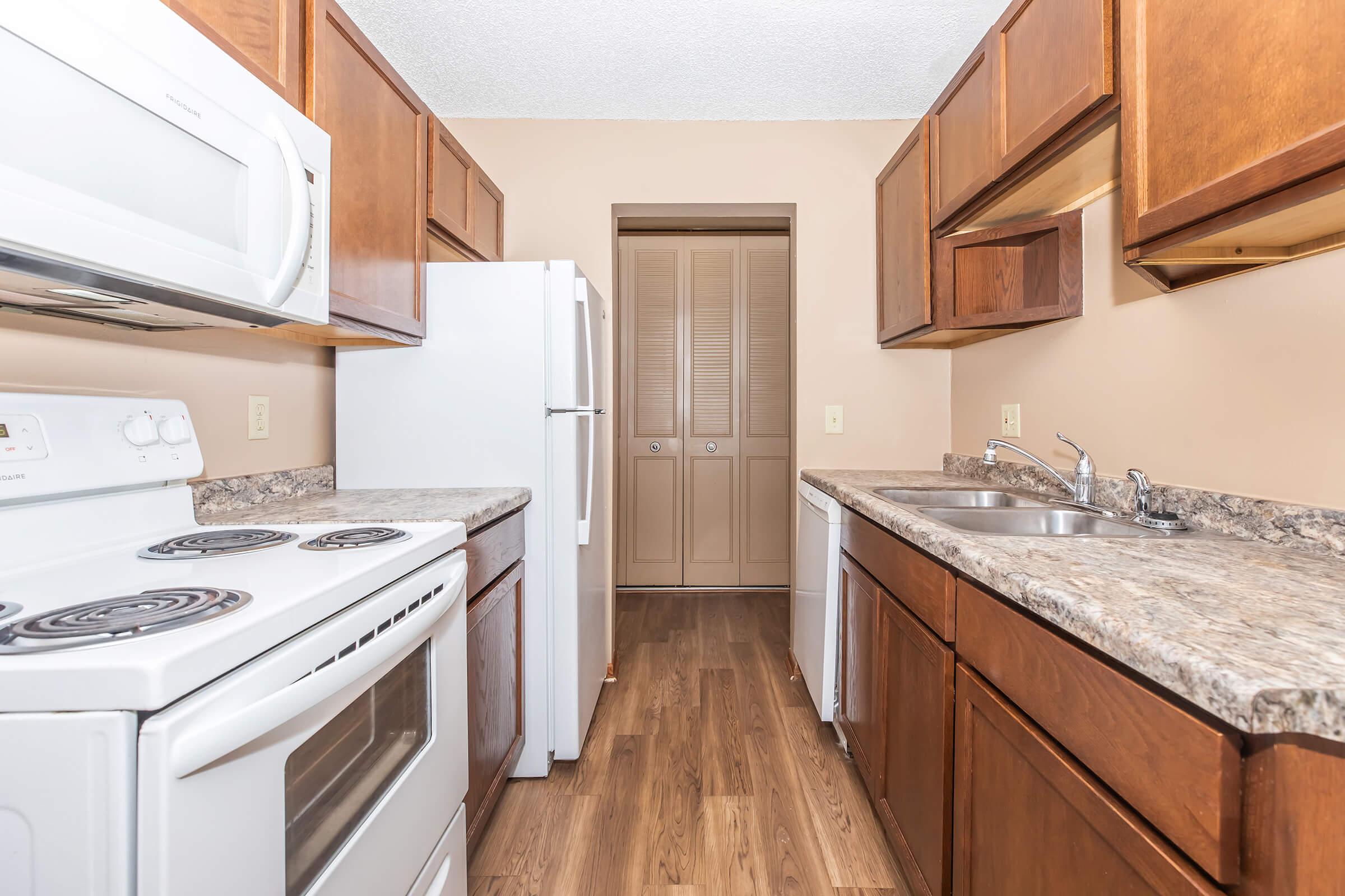 A well-lit kitchen featuring wooden cabinets, a white microwave, stove, and refrigerator. The countertops are made of a speckled material, and there are dual sinks. The floor is covered with laminate flooring, and a doorway leads to a storage area with closed closet doors.