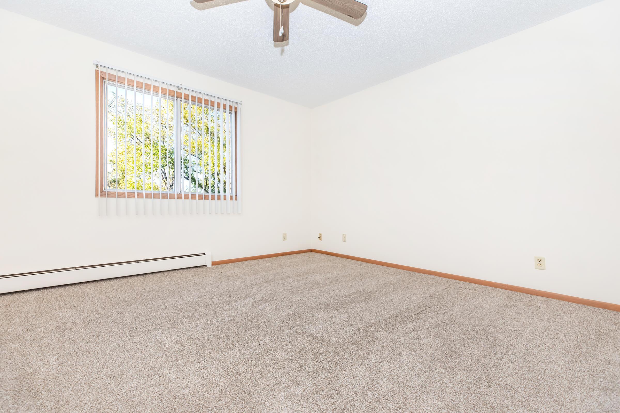 Empty room featuring beige carpet and white walls. A ceiling fan is mounted in the center, and a window with vertical blinds allows natural light to enter. The space is uncluttered, providing a bright and airy feel. No furniture is present.