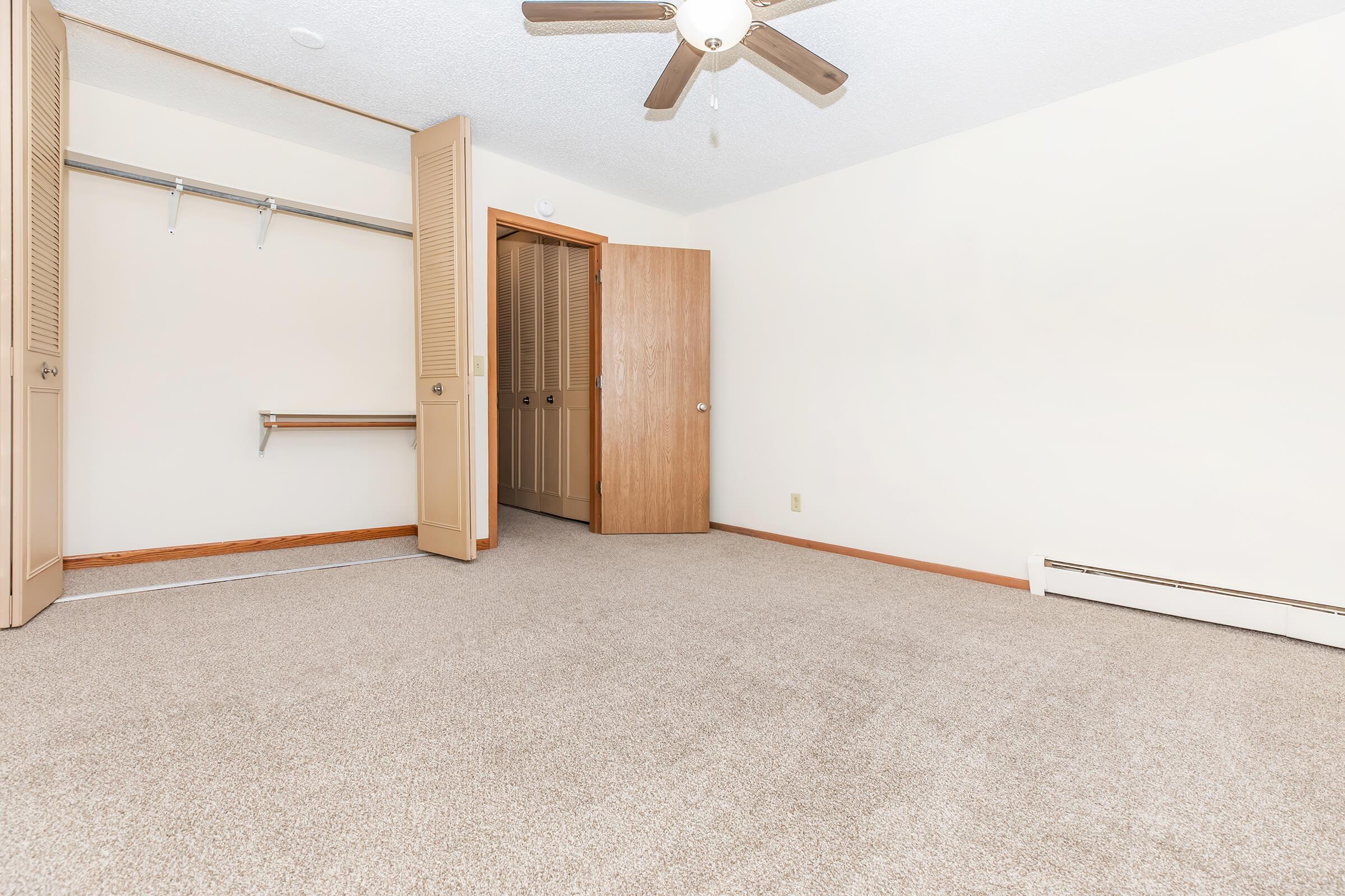 A spacious, empty room featuring light beige carpet, a ceiling fan, and a large closet with sliding doors. The walls are painted a soft cream color. There is an open door leading to another room, and the overall atmosphere is bright and uncluttered, providing a blank canvas for personal decoration.