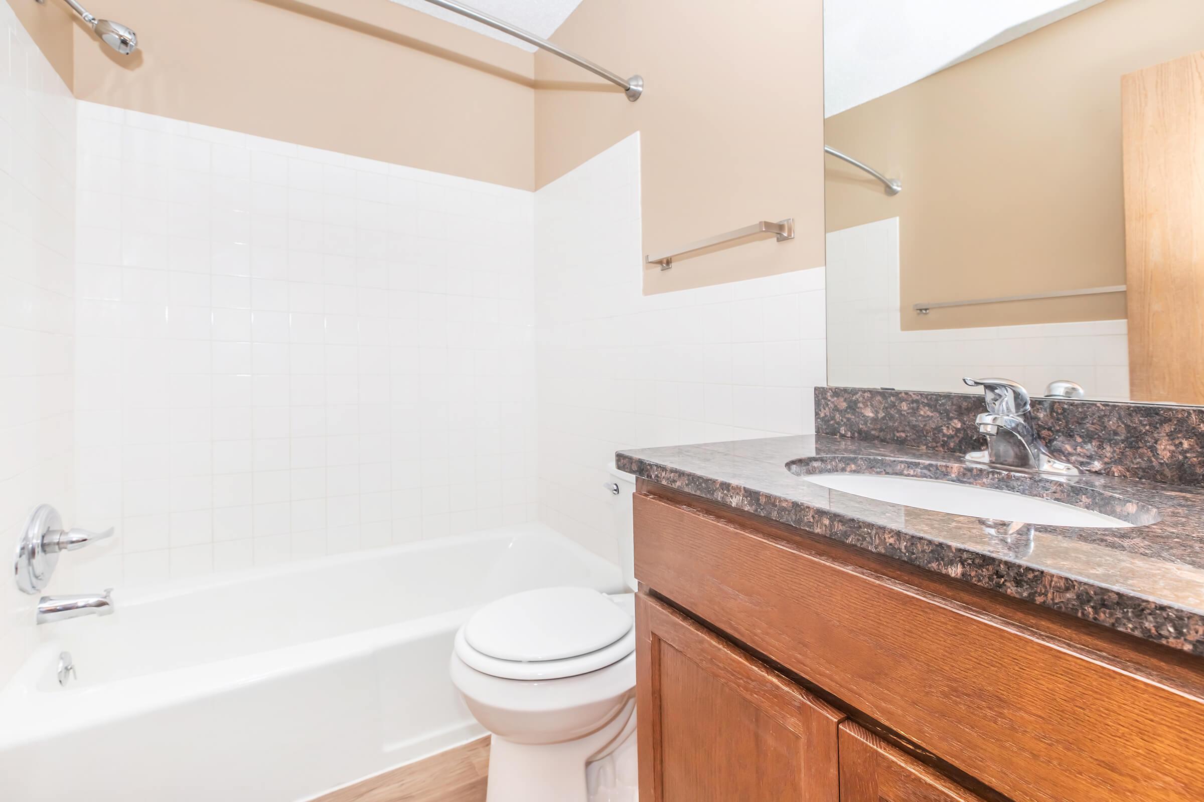 A clean, modern bathroom featuring a white bathtub, a toilet, and a countertop with a sink. The walls are painted a light beige, and there's a mirror above the sink. The floor appears to be a light wood finish, creating a bright and inviting space.