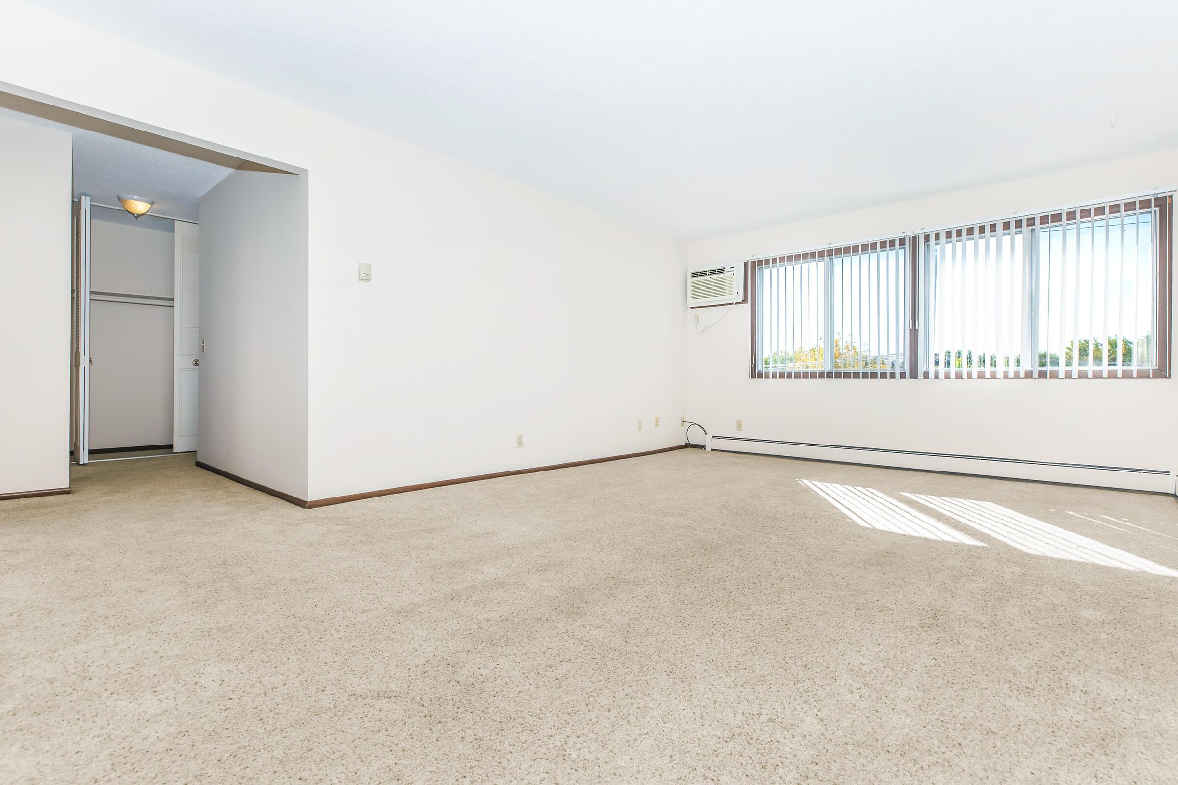 An empty, well-lit room with beige carpet and white walls. There is a window on the right with horizontal blinds allowing natural light to enter. An air conditioning unit is visible near the window. A doorway leads to another space on the left. The overall atmosphere is clean and minimalist.