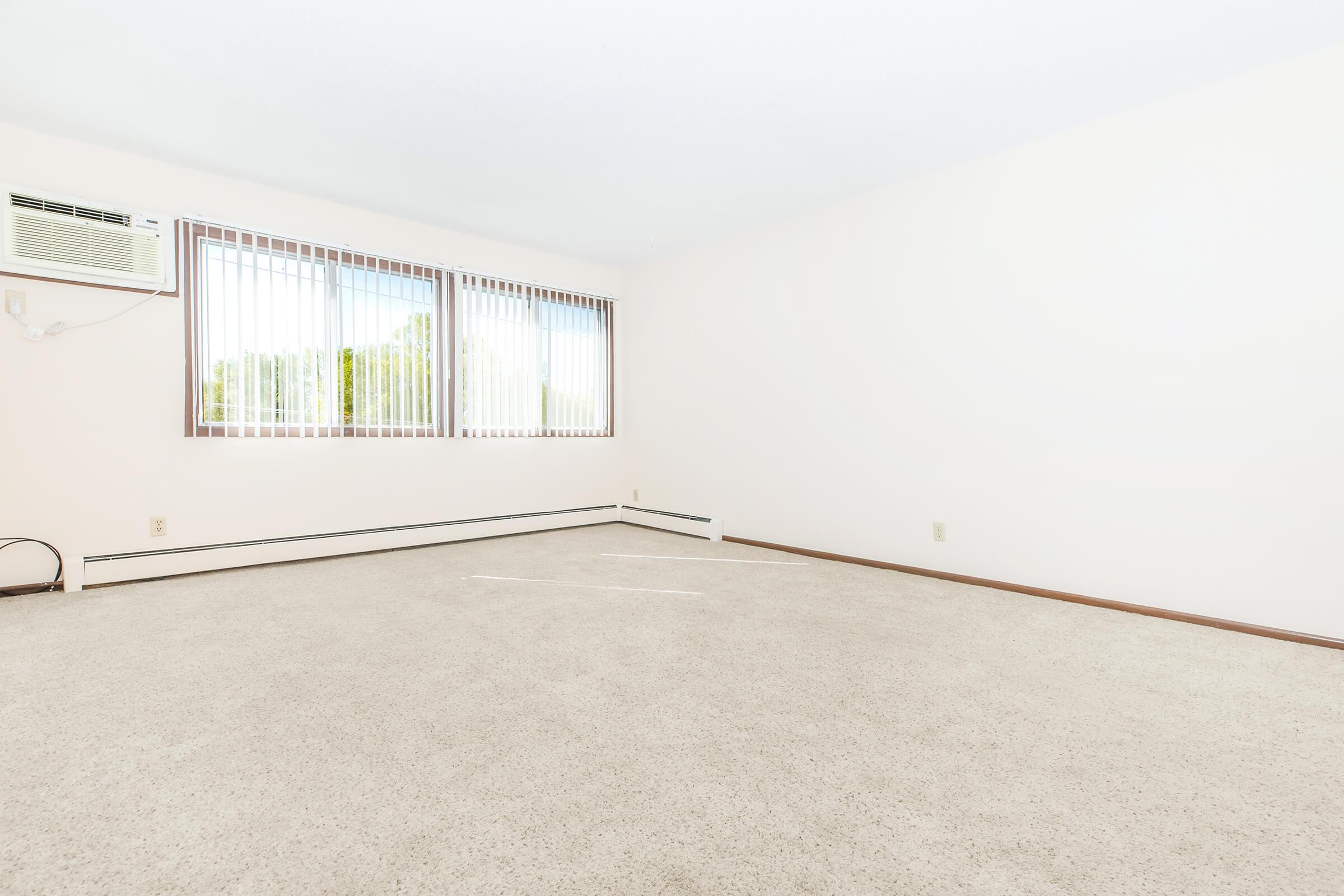 A spacious, empty room with light-colored walls and carpet. There are large windows with blinds allowing natural light to enter. An air conditioning unit is mounted on the wall, and there are no furniture or decorations present, creating a blank canvas for potential customization.