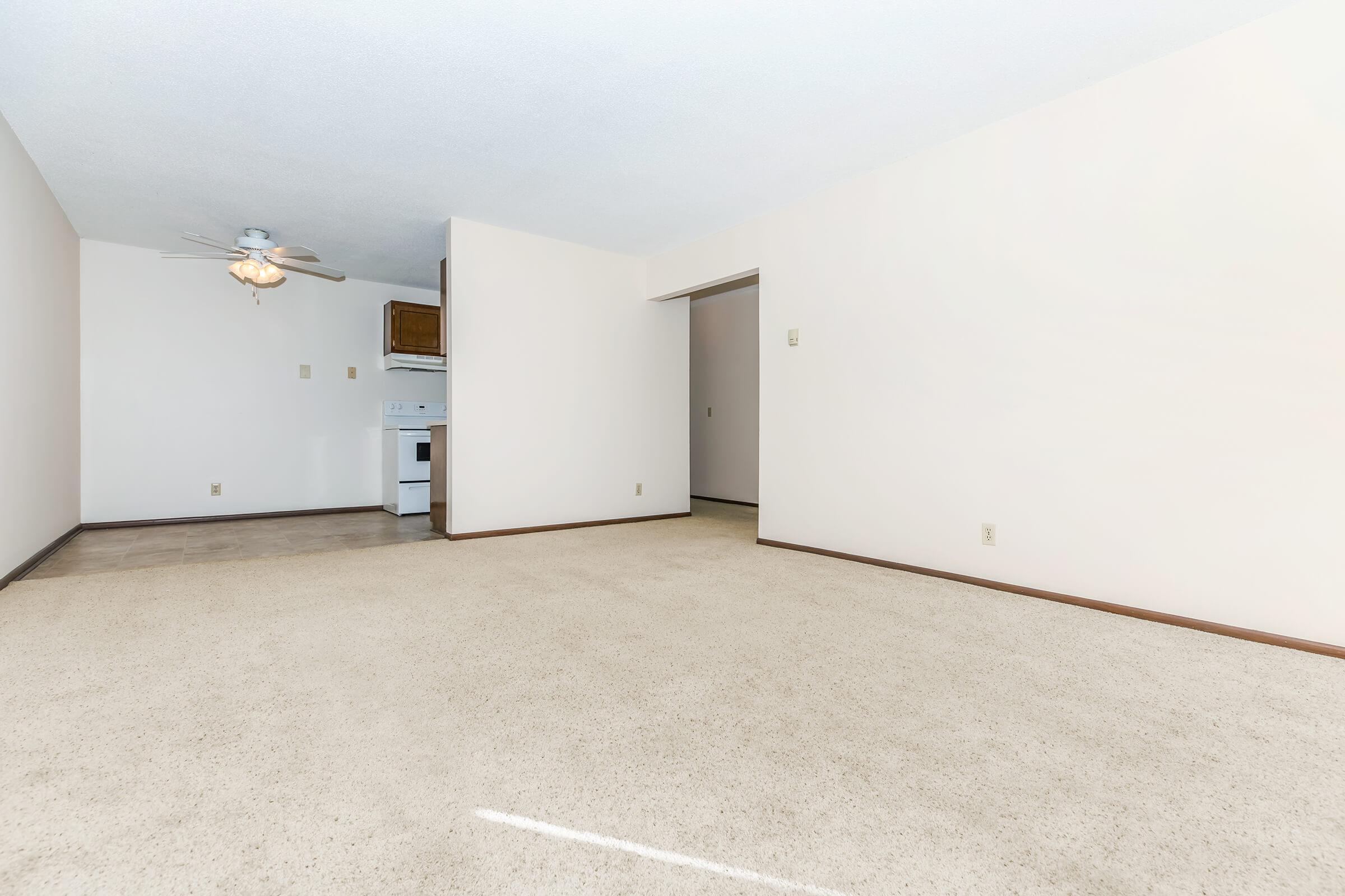 A spacious, empty room with beige carpet and light-colored walls. A ceiling fan is mounted above, and there's a small kitchen area visible with a stove and cabinets. The space has a bright, airy feel with natural light. No furniture or decorations are present.