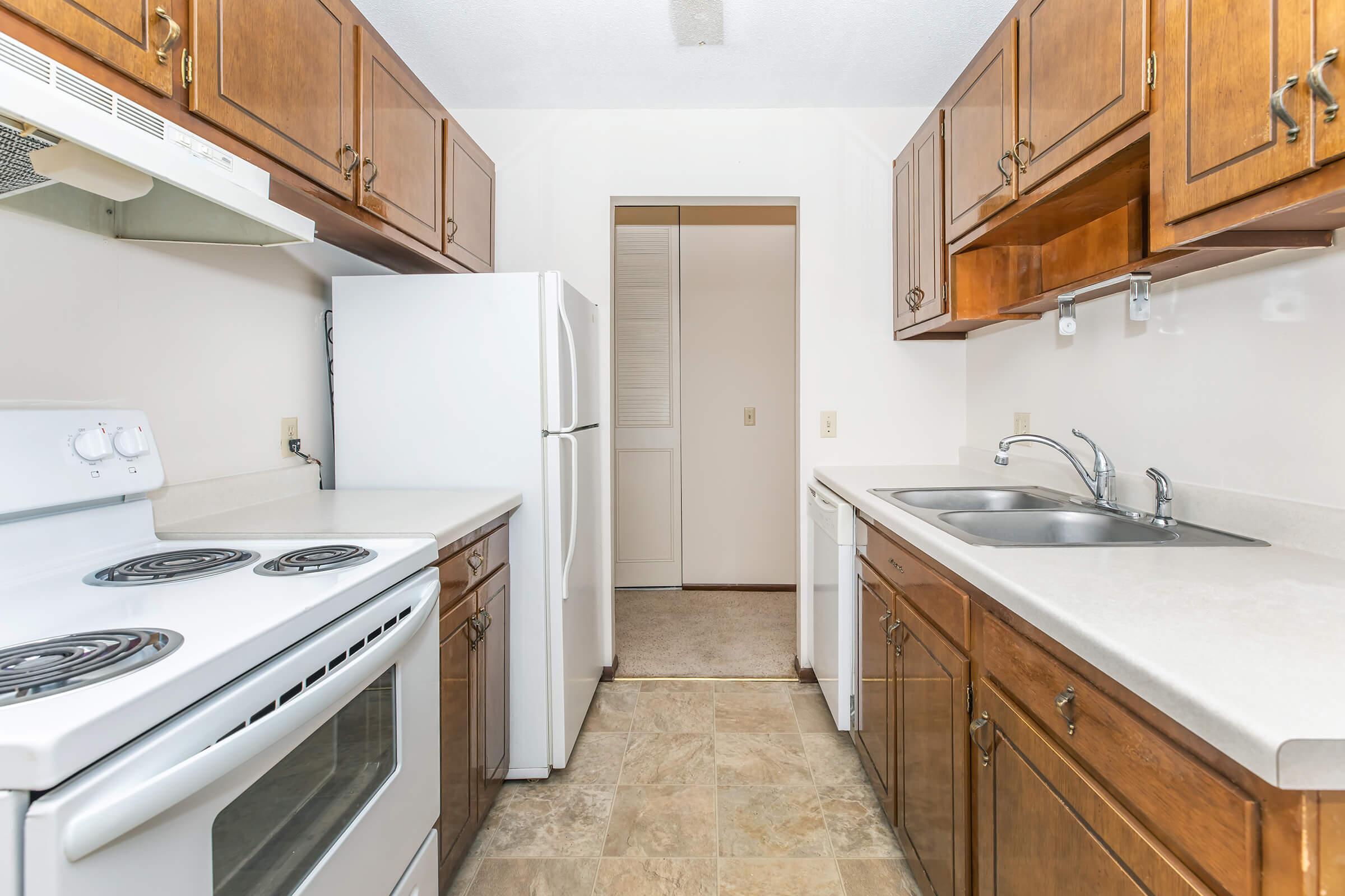 A modern, clean kitchen with wooden cabinets, a white stove, a refrigerator, and a double sink. The countertops are light-colored, and the flooring has a tile pattern. An open doorway leads to another room, suggesting a spacious layout. The overall atmosphere is bright and inviting.