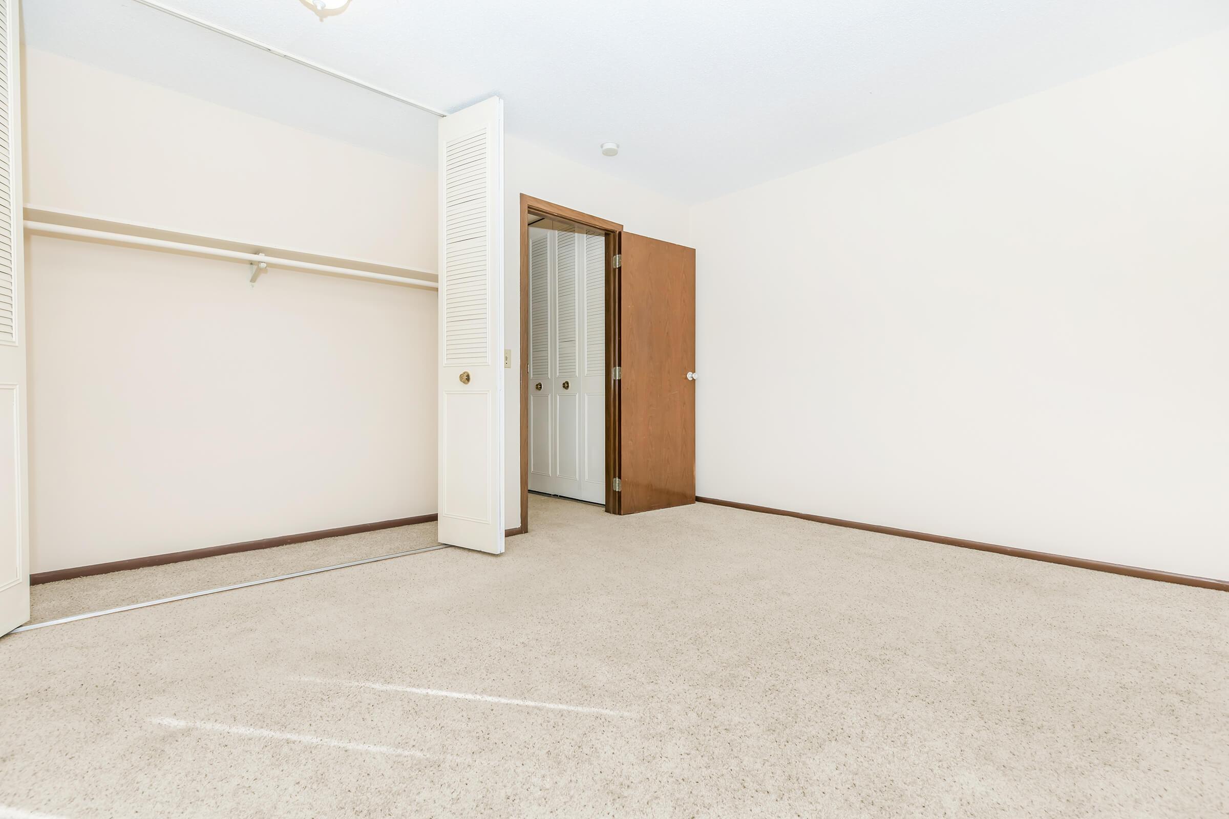 An empty room with beige walls and carpeting. A closet with sliding doors is on the left, while a closed wooden door is on the right. The space is well-lit, with sunlight streaming in, creating a bright and open atmosphere. No furniture or decorations are present, emphasizing the room's emptiness.