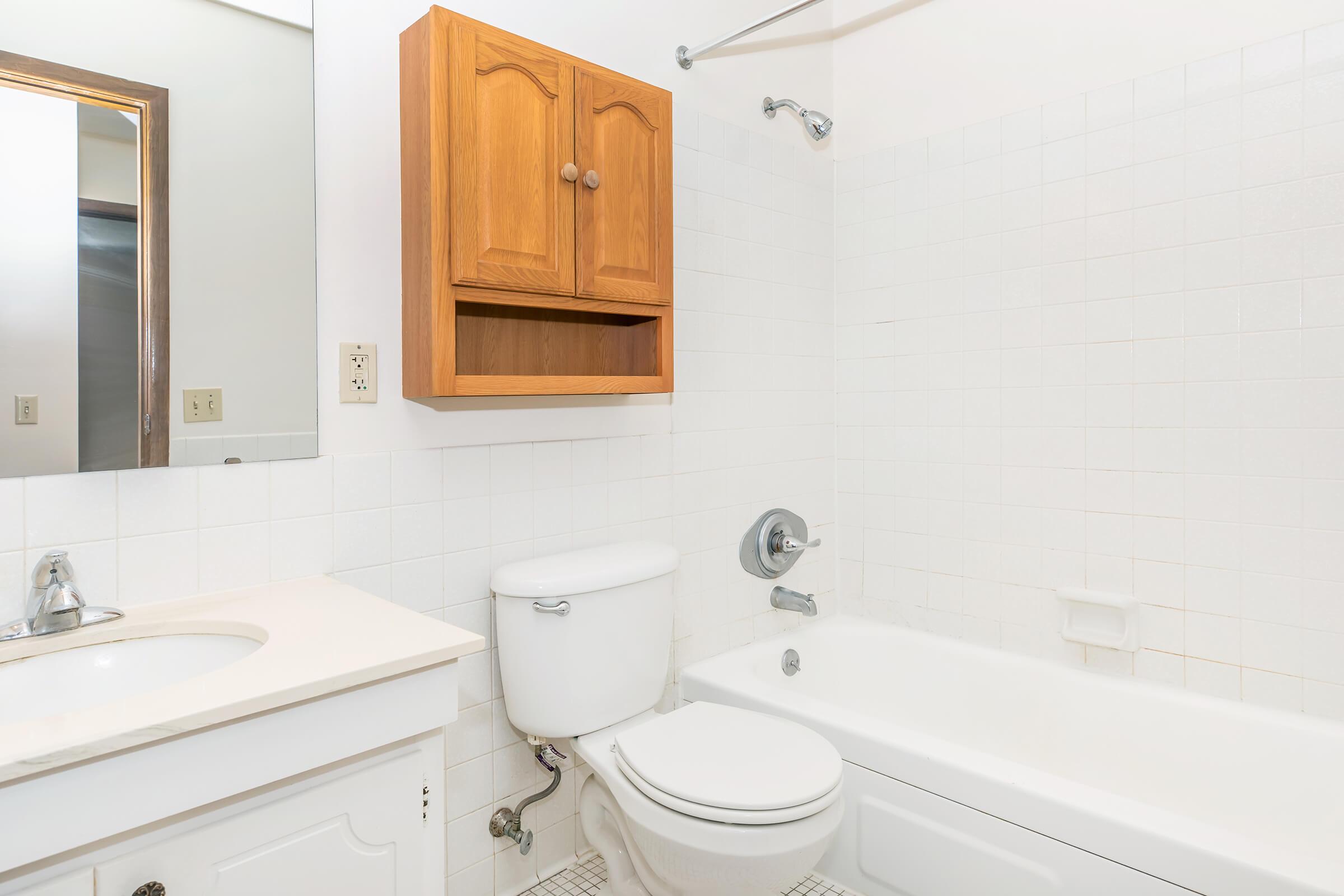 A clean and simple bathroom featuring a white bathtub, a toilet, a sink with a countertop, and a wooden cabinet above the sink. The walls and tiles are mostly white, with a mirror above the sink and a showerhead mounted on the wall. Natural light is present from a nearby window.