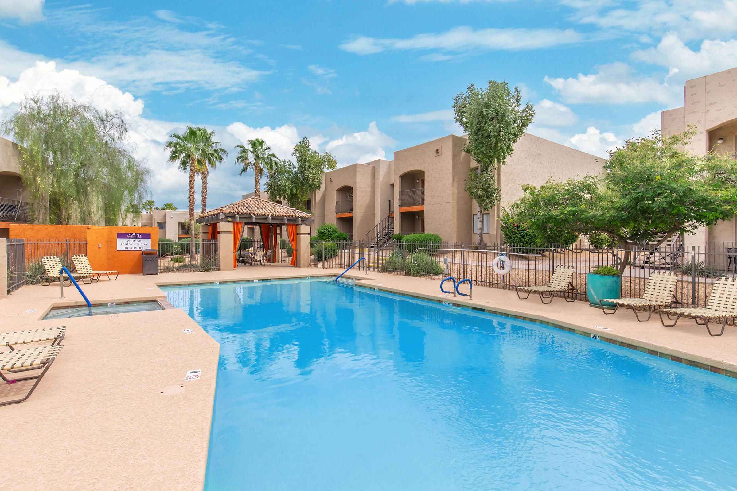 A beautifully designed outdoor swimming pool surrounded by lounge chairs and palm trees, with a gazebo nearby. The pool area features clear blue water under a bright sky, complemented by modern apartment buildings in the background.