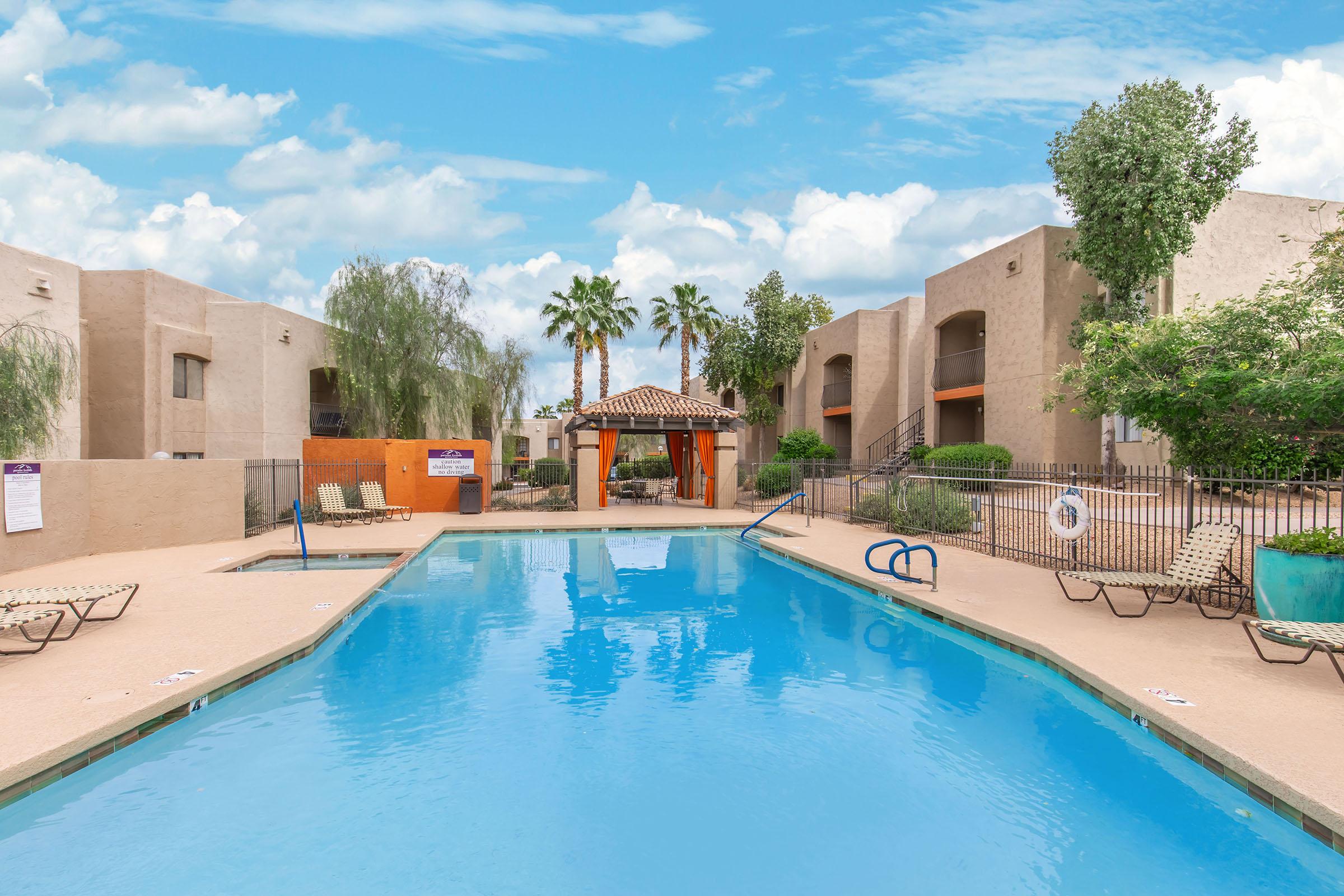 A clear blue swimming pool surrounded by lounge chairs and palm trees, with a gazebo in the background. The surrounding buildings are light-colored with a desert aesthetic. The sky above is bright blue with fluffy white clouds, creating a relaxing outdoor atmosphere.