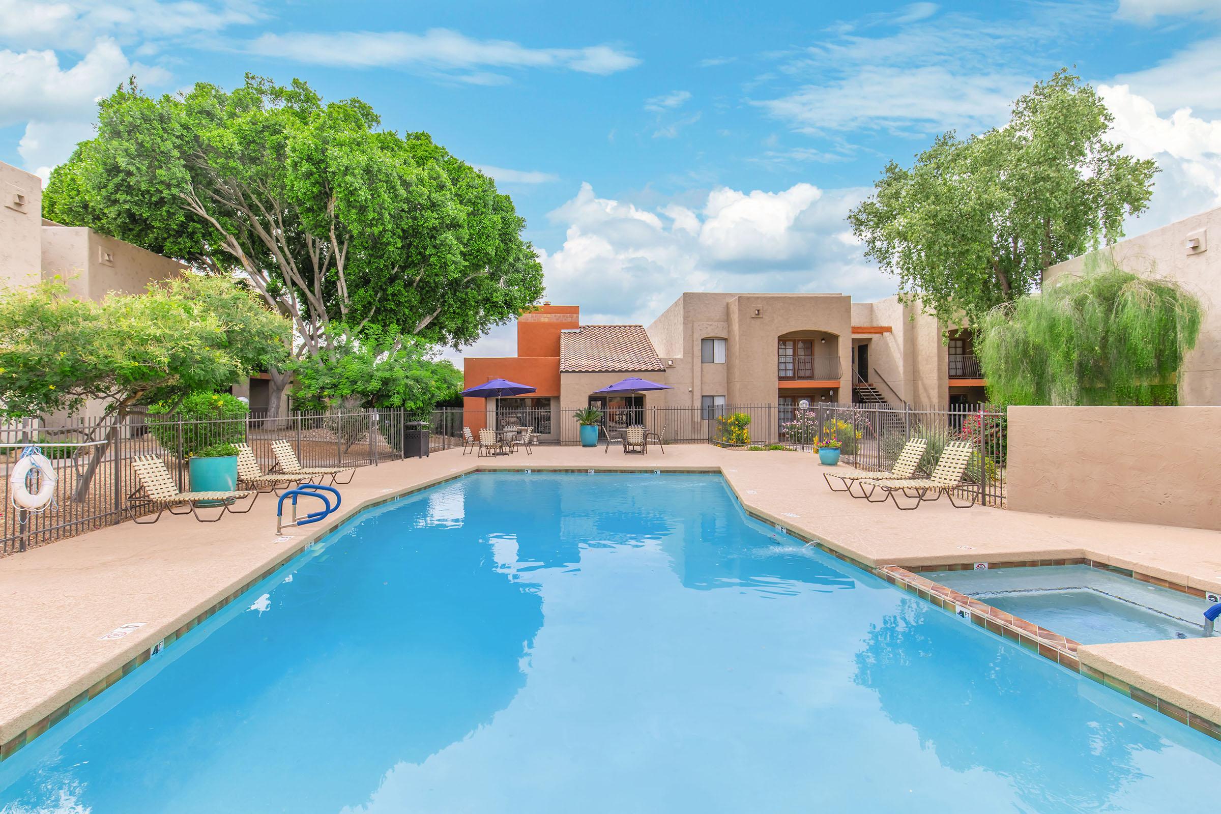 A bright outdoor pool surrounded by lounge chairs and vibrant trees. In the background, there are buildings with balconies and patio areas. The sky is clear with fluffy clouds, creating a relaxing and inviting atmosphere for leisure activities.