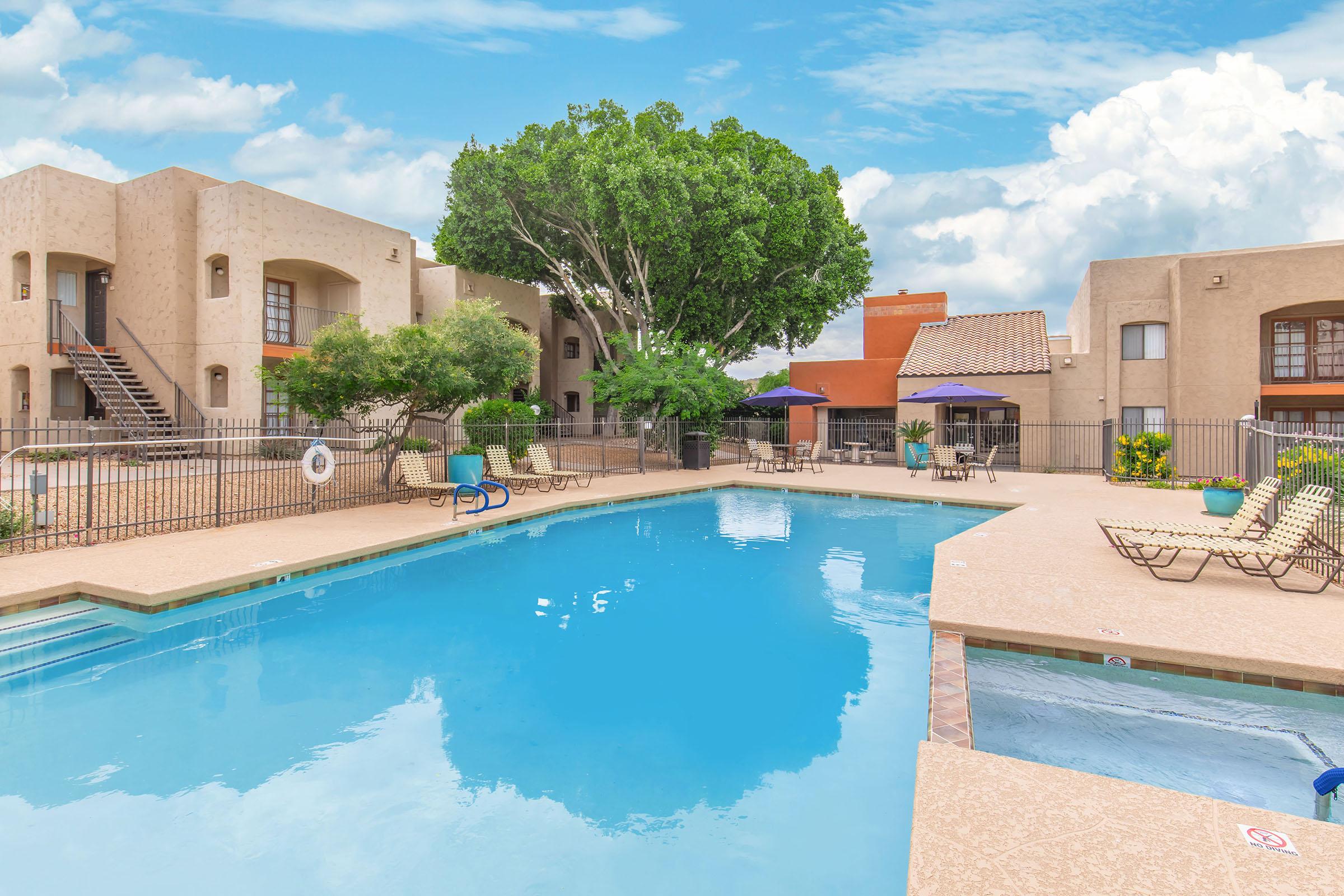 A clear swimming pool surrounded by lounge chairs, trees, and an apartment complex. The sky is partly cloudy, creating a bright and inviting atmosphere for relaxation.