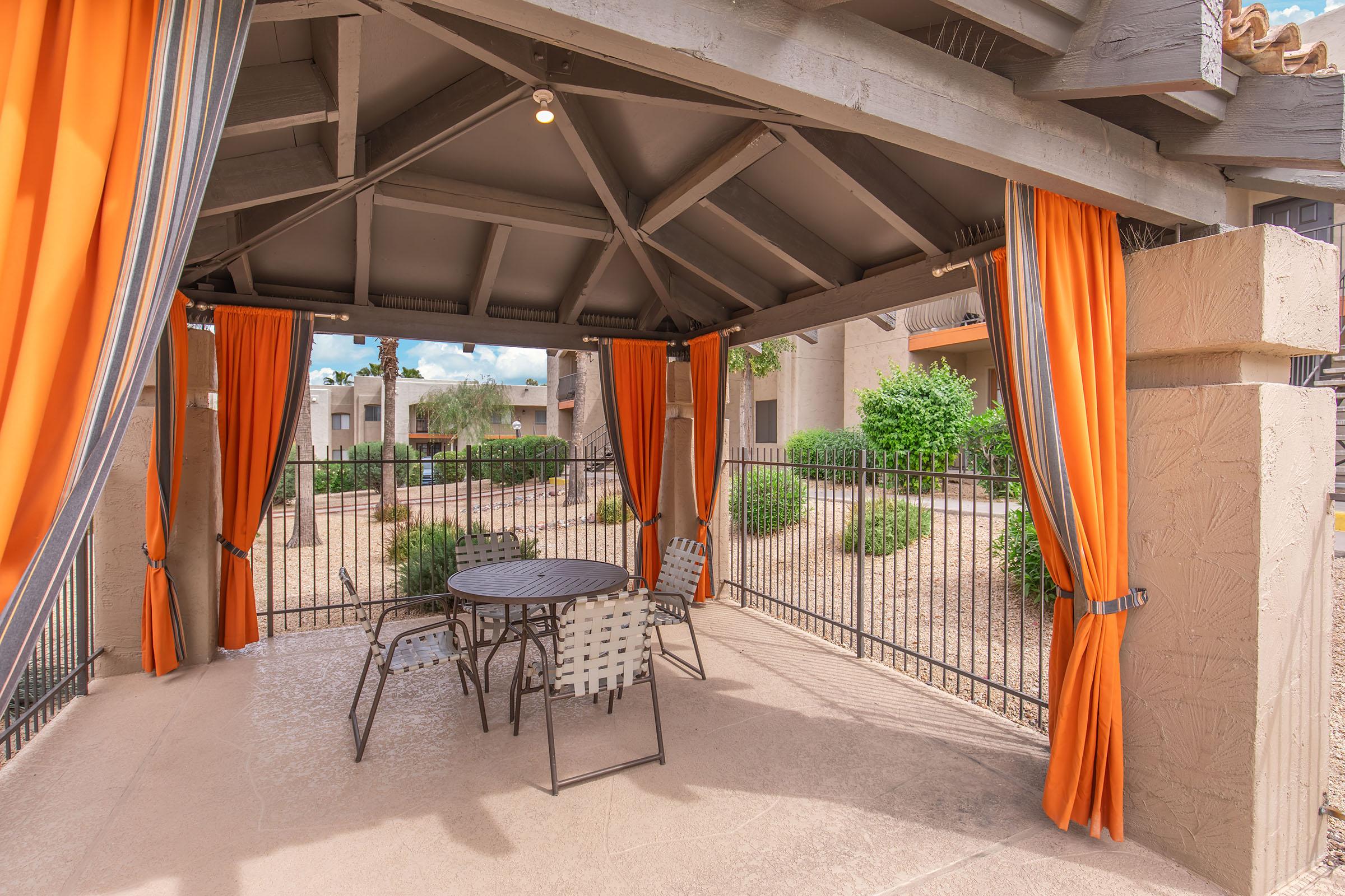A shaded outdoor gazebo with orange curtains, featuring a round table and metal chairs. Lush greenery and a fence are visible in the background, under a blue sky with clouds. The setting provides a comfortable space for relaxation or social gatherings.