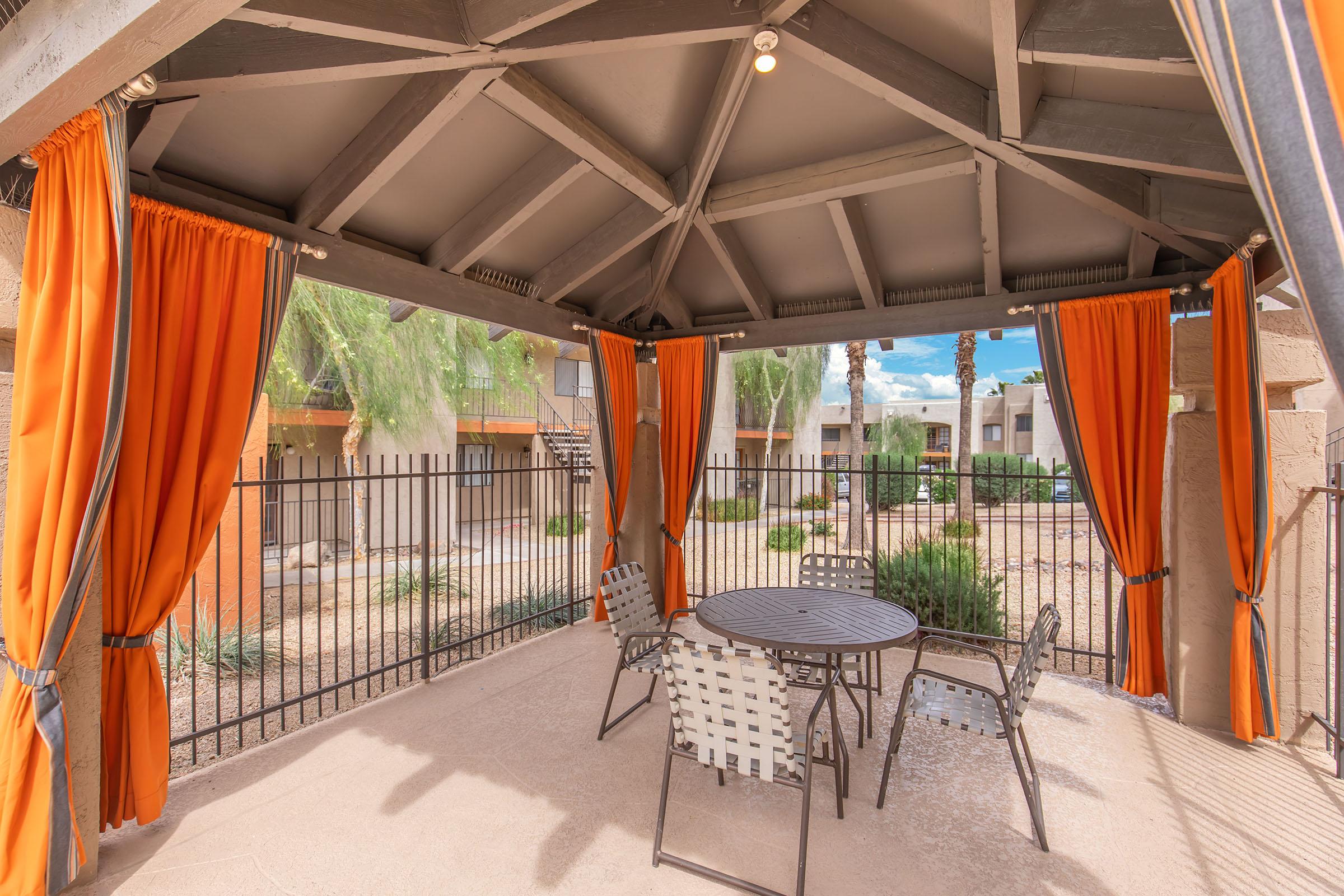 A shaded outdoor gazebo featuring a round table and four chairs. The gazebo is adorned with orange curtains, and the background shows a landscaped area with greenery and residential buildings. The setting appears inviting and suited for relaxation or social gatherings.