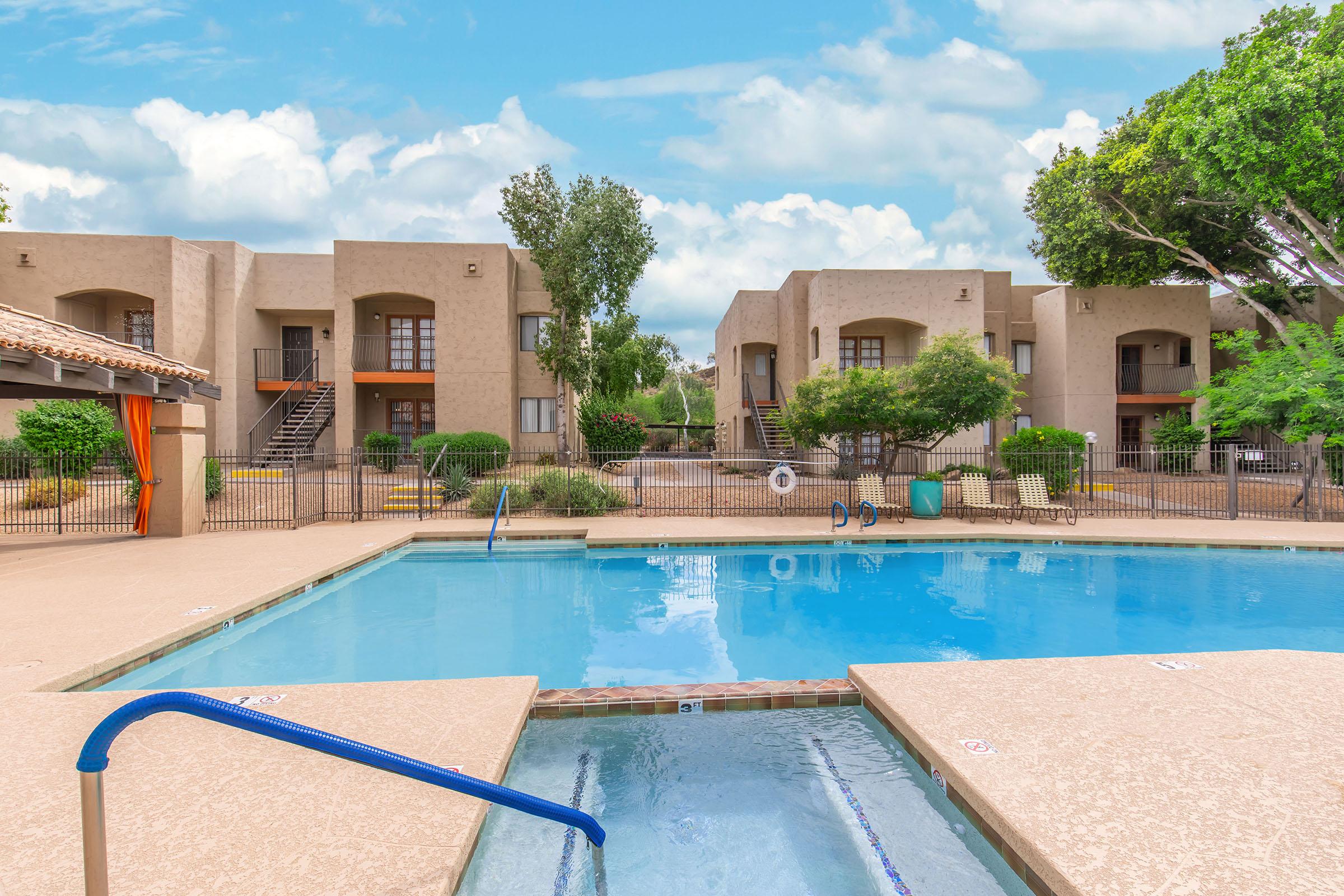 A clear blue swimming pool surrounded by a patio with lounge chairs. Apartment buildings with balconies are visible in the background, along with green shrubs and trees. The sky is partly cloudy, creating a serene atmosphere in a residential area.