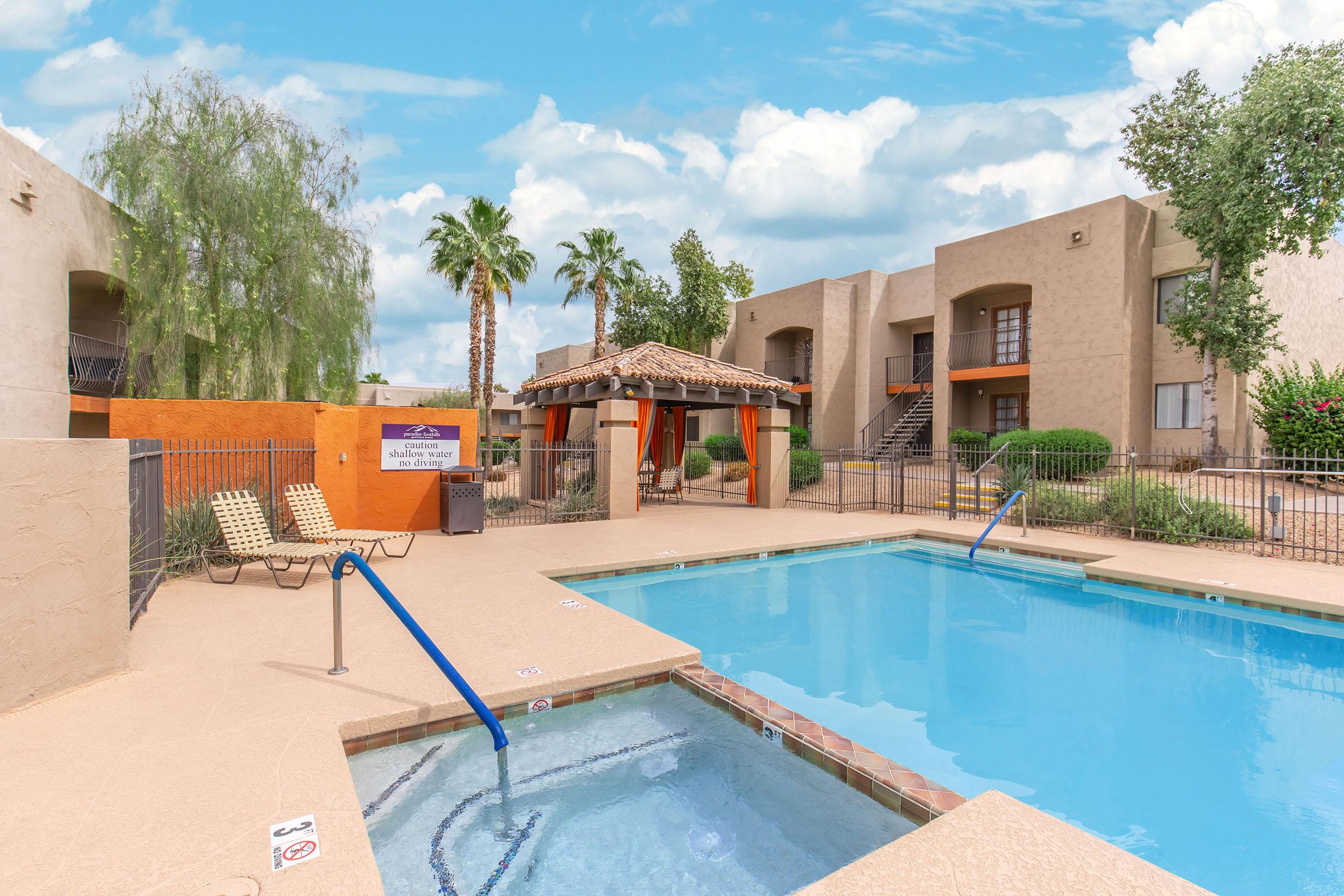 A well-maintained outdoor pool area featuring a swimming pool and a hot tub. Surrounding the pool are lounge chairs and a shaded cabana. The background includes palm trees and modern apartment buildings under a partly cloudy sky.
