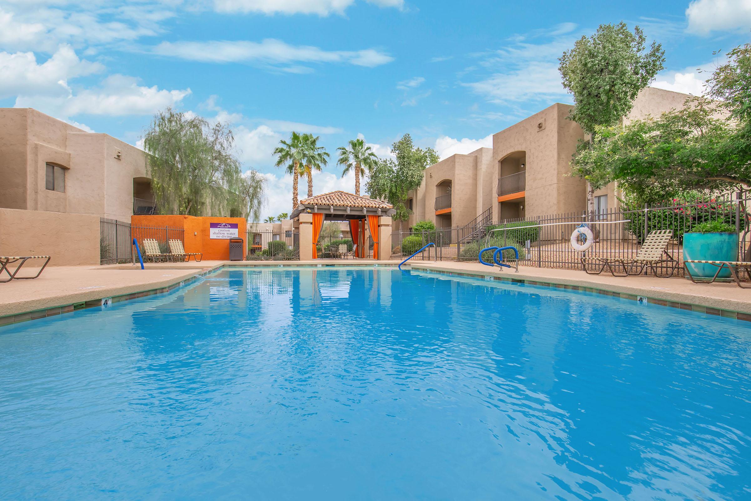 A large swimming pool with crystal-clear blue water, surrounded by lounge chairs. In the background, palm trees and landscaped greenery add to the ambiance. On one side, there is a covered seating area with a tiled roof, and residential buildings frame the scene under a bright blue sky.