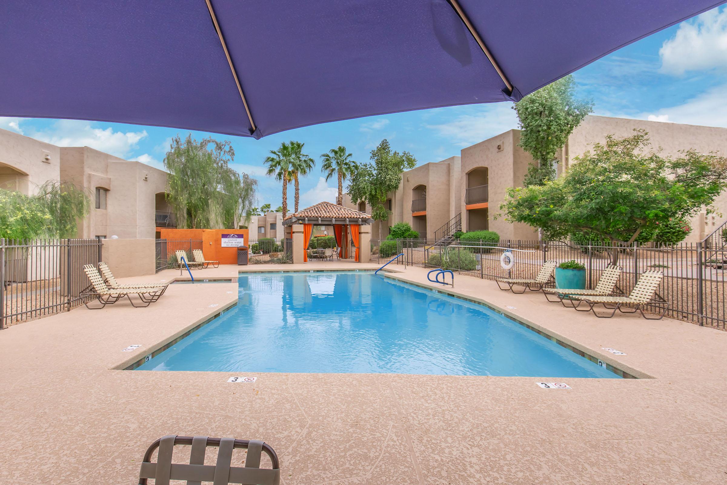 A serene swimming pool surrounded by lounge chairs, with an umbrella providing shade. Lush palm trees and colorful landscaping frame the area, along with buildings in a desert-style architecture. The clear blue water reflects the sky, creating a relaxing atmosphere.
