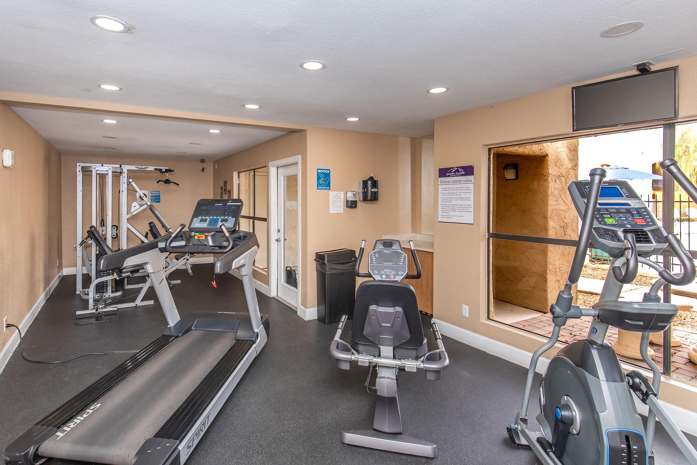 A modern gym interior featuring various exercise equipment, including treadmills, an elliptical machine, and a weight machine. The walls are painted in a light color, and there's a large window allowing natural light. A flat-screen TV is mounted on the wall, enhancing the workout environment.