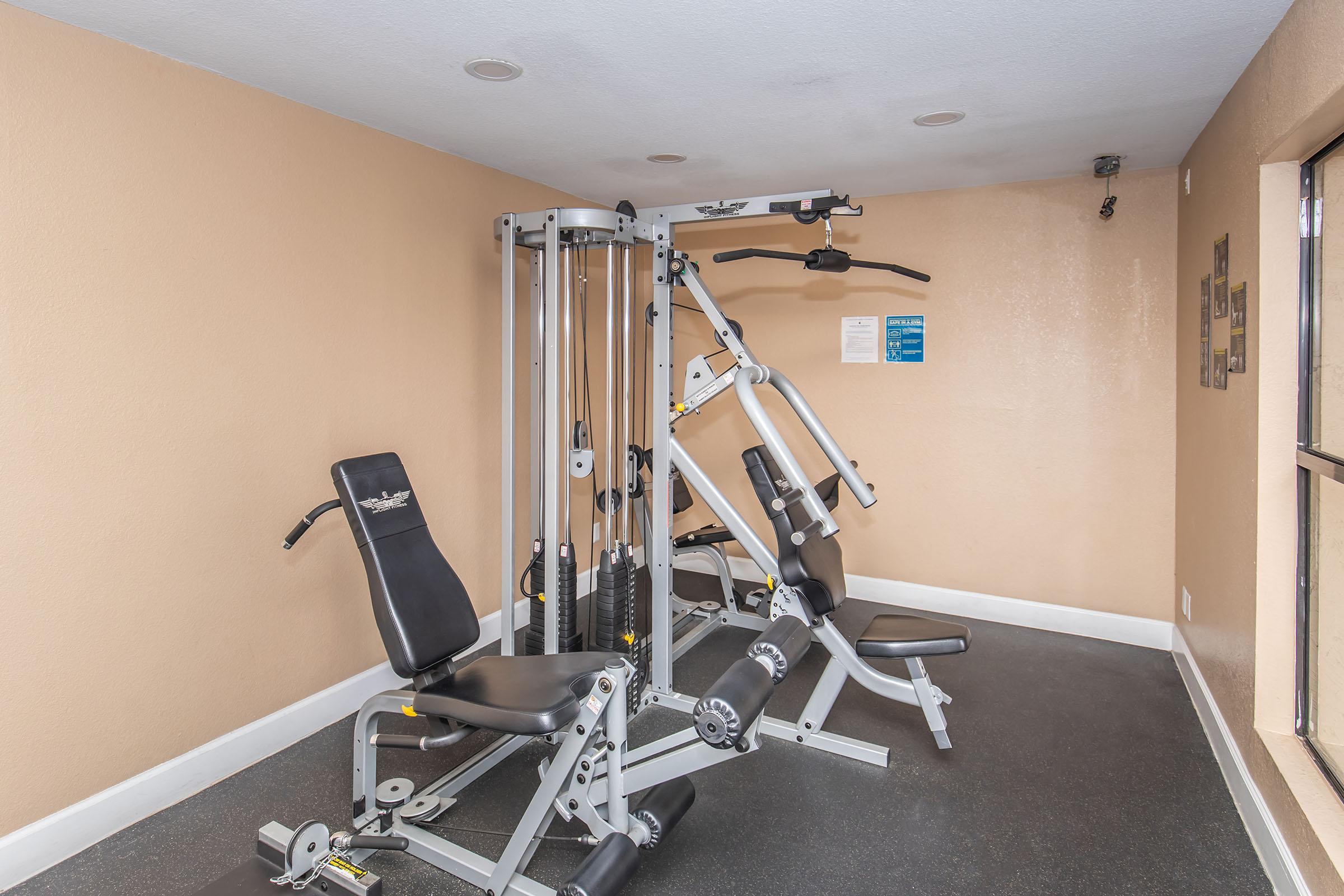 A small gym area featuring a multi-gym machine and a seated weight bench. The walls are painted in a light peach color, and there are circular ceiling lights. A window lets in natural light, and there is a blue informational sign on the wall. The flooring is dark and carpeted.