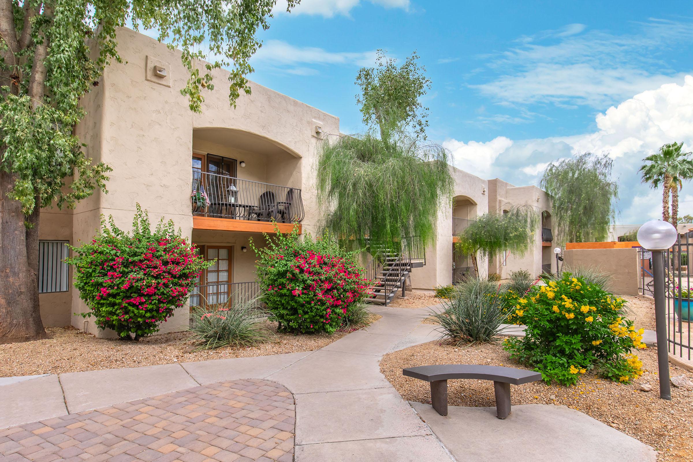View of a well-maintained apartment complex with beige stucco buildings, surrounded by vibrant flowering shrubs and palm trees. A paved pathway winds through the landscape, leading to the entrances of the apartments. Clear blue sky above adds to the serene atmosphere of the environment.