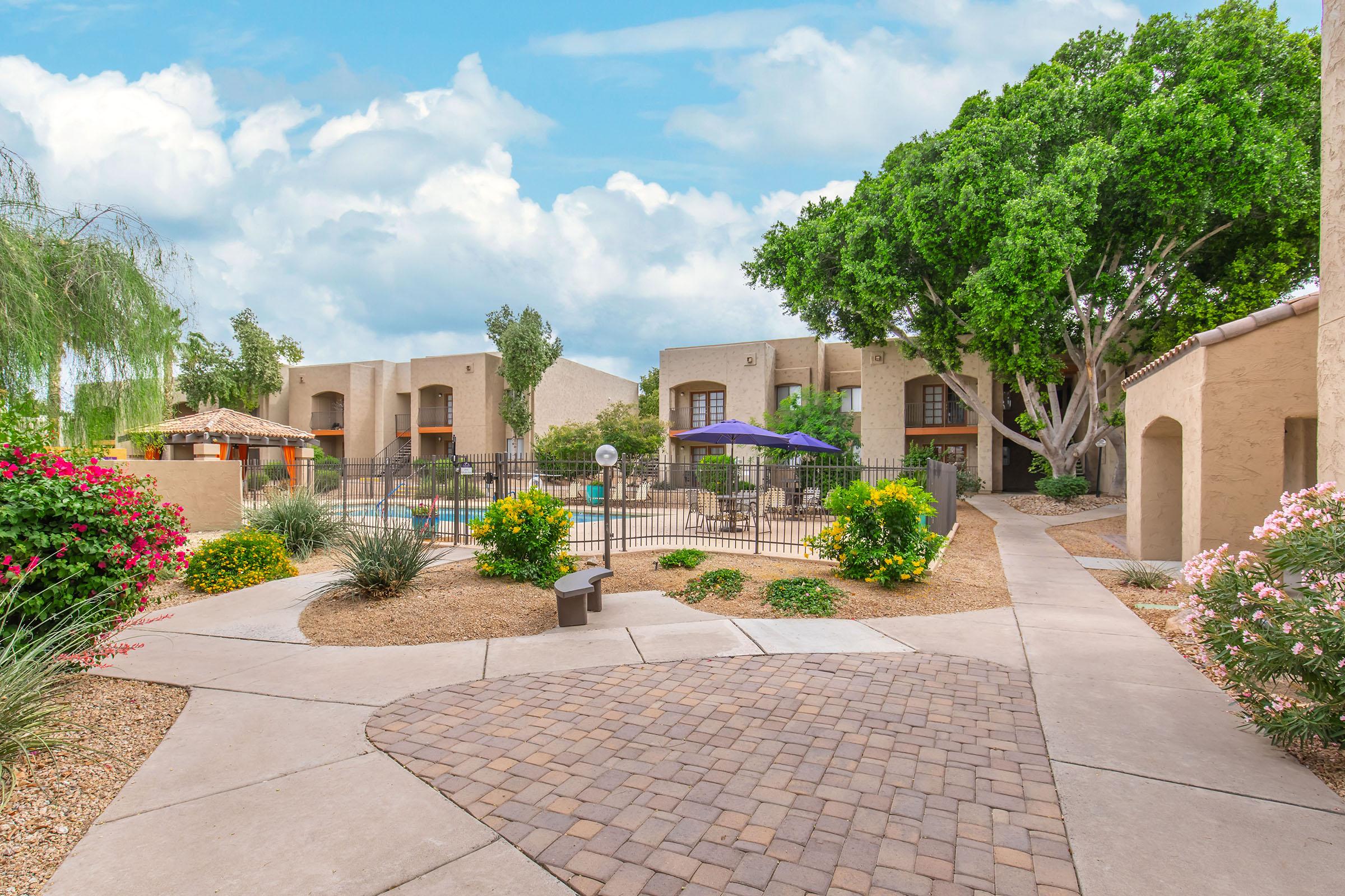 A peaceful courtyard featuring a paved walkway, vibrant flower bushes, and a swimming pool surrounded by fences and patio areas. The background includes two-story apartment buildings with balconies, under a partly cloudy sky. Green trees provide shade, creating a serene atmosphere.