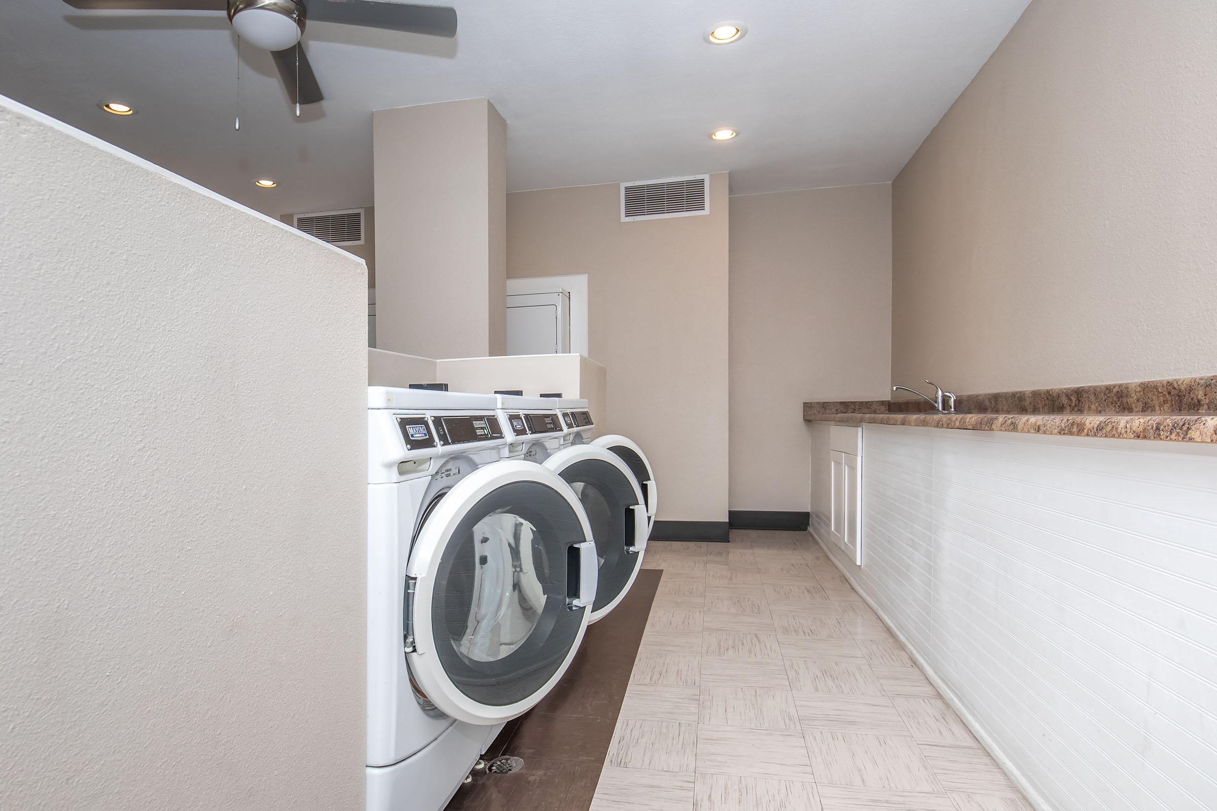 A laundry room featuring two white washers and dryers positioned against a light-colored wall. There is a countertop space next to the appliances and a fan on the ceiling providing ventilation. The floor is tiled, and the overall space is well-lit.