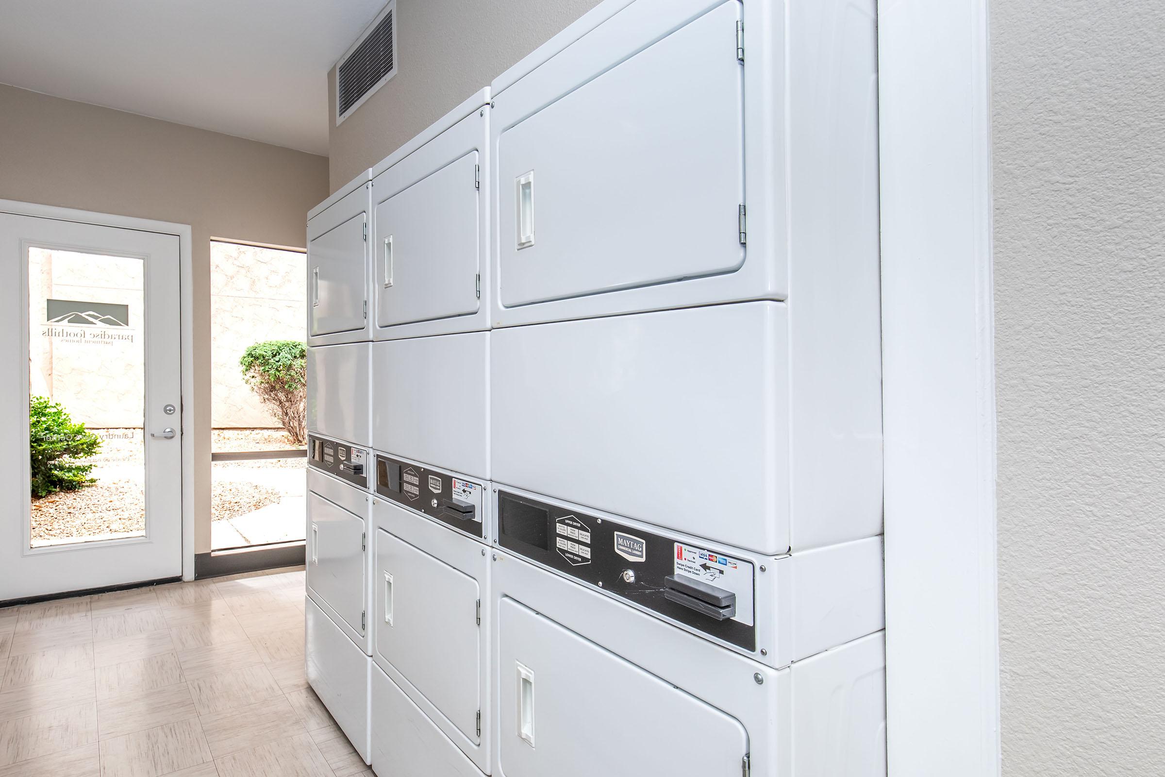 Row of white coin-operated laundry machines against a beige wall, with a partially visible door leading outside. The area is well-lit, and there are plants outside the door, suggesting an entrance to a laundry facility.