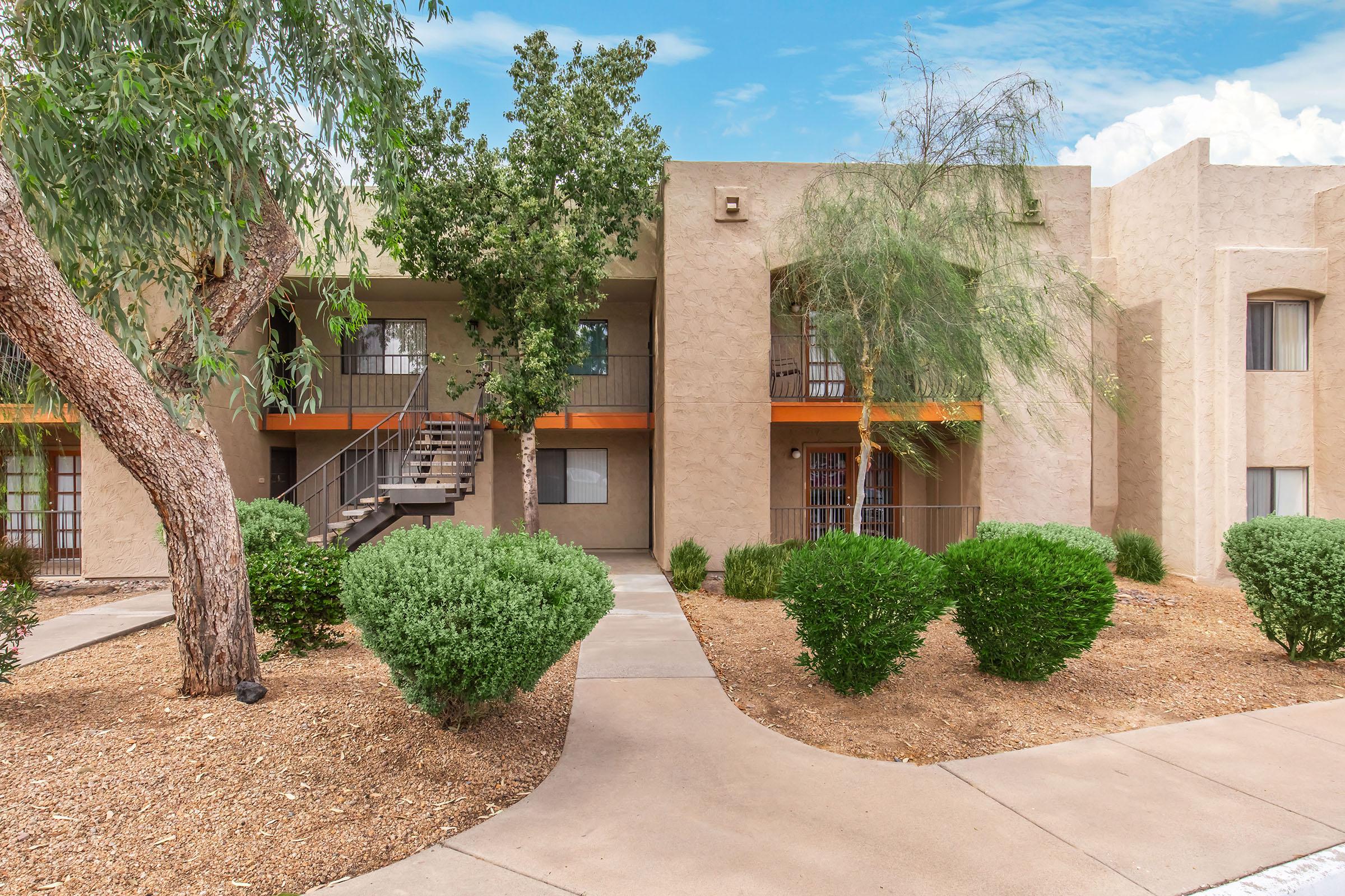 A well-maintained apartment complex featuring two-story buildings with a landscaped pathway. Lush green shrubs and trees surround the area, providing a welcoming atmosphere. The buildings have beige exteriors with balconies and staircases, under a clear blue sky.