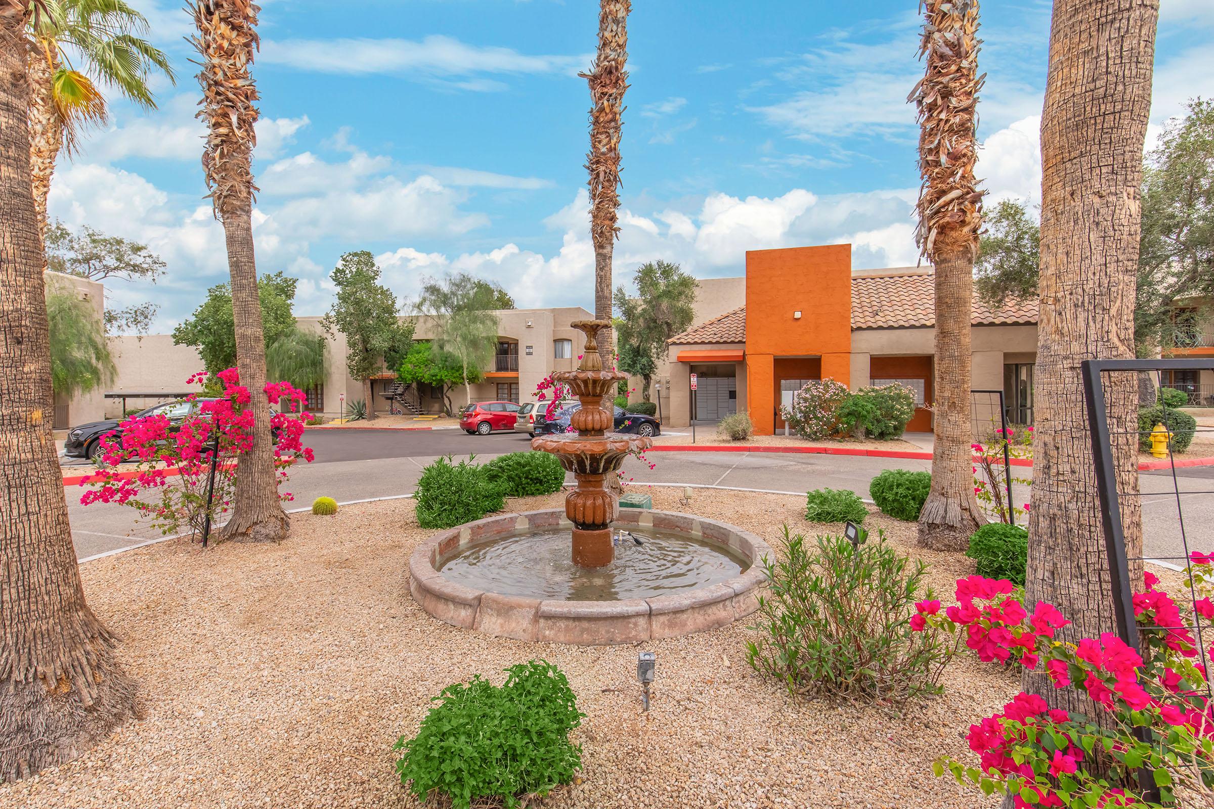A peaceful courtyard featuring a decorative stone fountain surrounded by vibrant pink bougainvillea flowers. Palm trees line the area, complementing the tranquil atmosphere. In the background, there are buildings with earthy tones and a clear blue sky overhead.