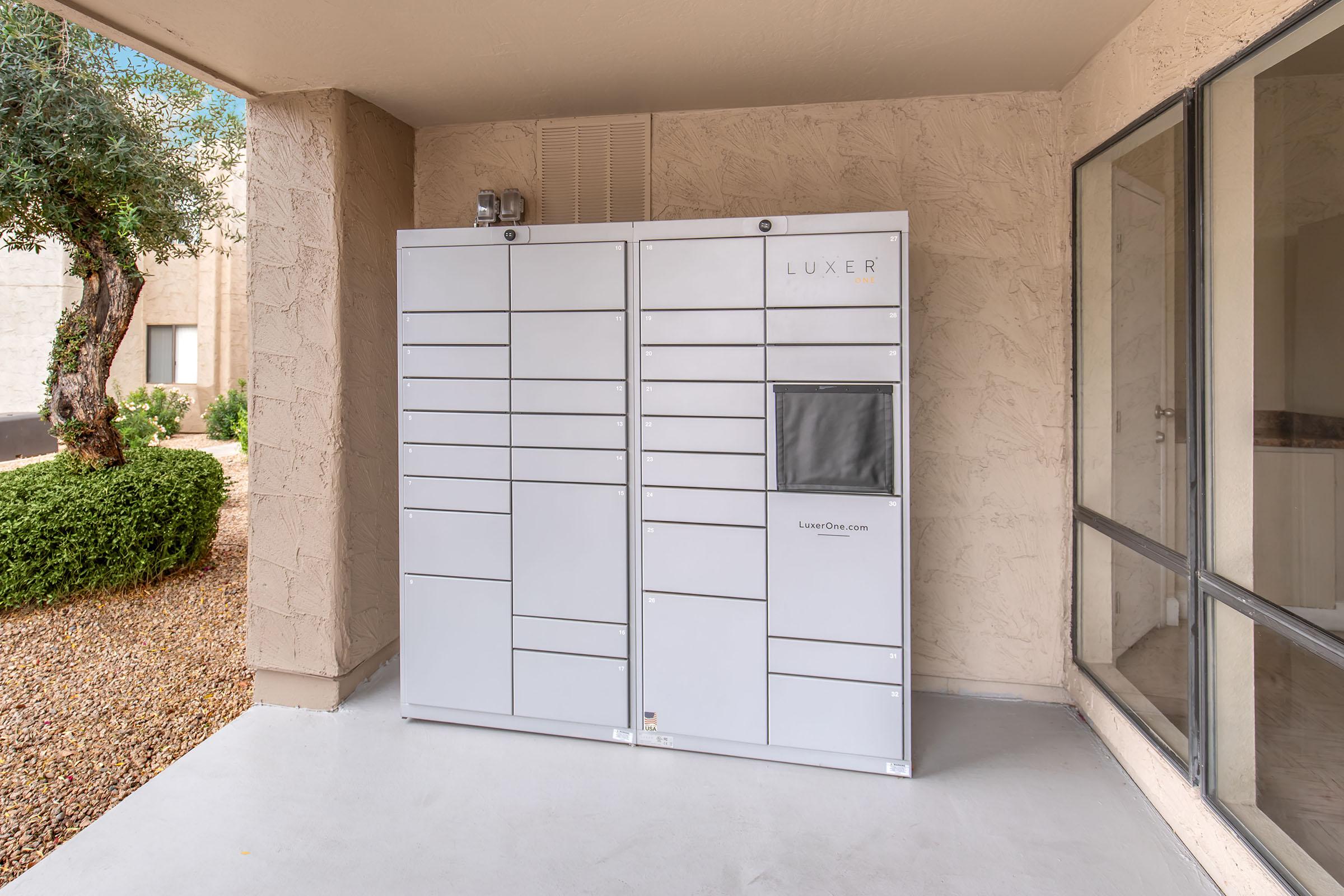 A white outdoor package delivery locker system placed on a porch, featuring multiple compartments and a digital screen. Surrounding the locker are desert landscaping elements, including small bushes and gravel, with a view of a building in the background. The overall setting is well-lit and tidy.