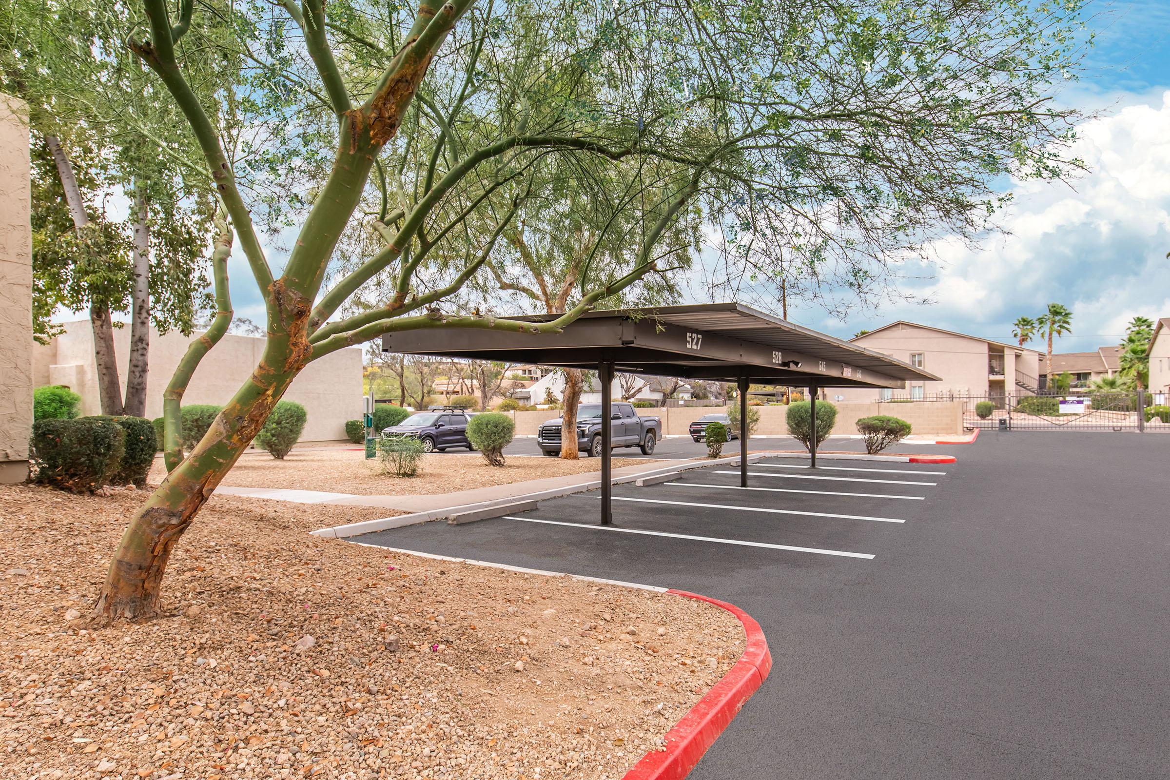 A parking lot with covered parking spaces and well-maintained landscaping, featuring a tree and shrubs. In the background, there are residential buildings and a partially cloudy sky. The pavement is freshly laid, and there are a few parked cars visible.