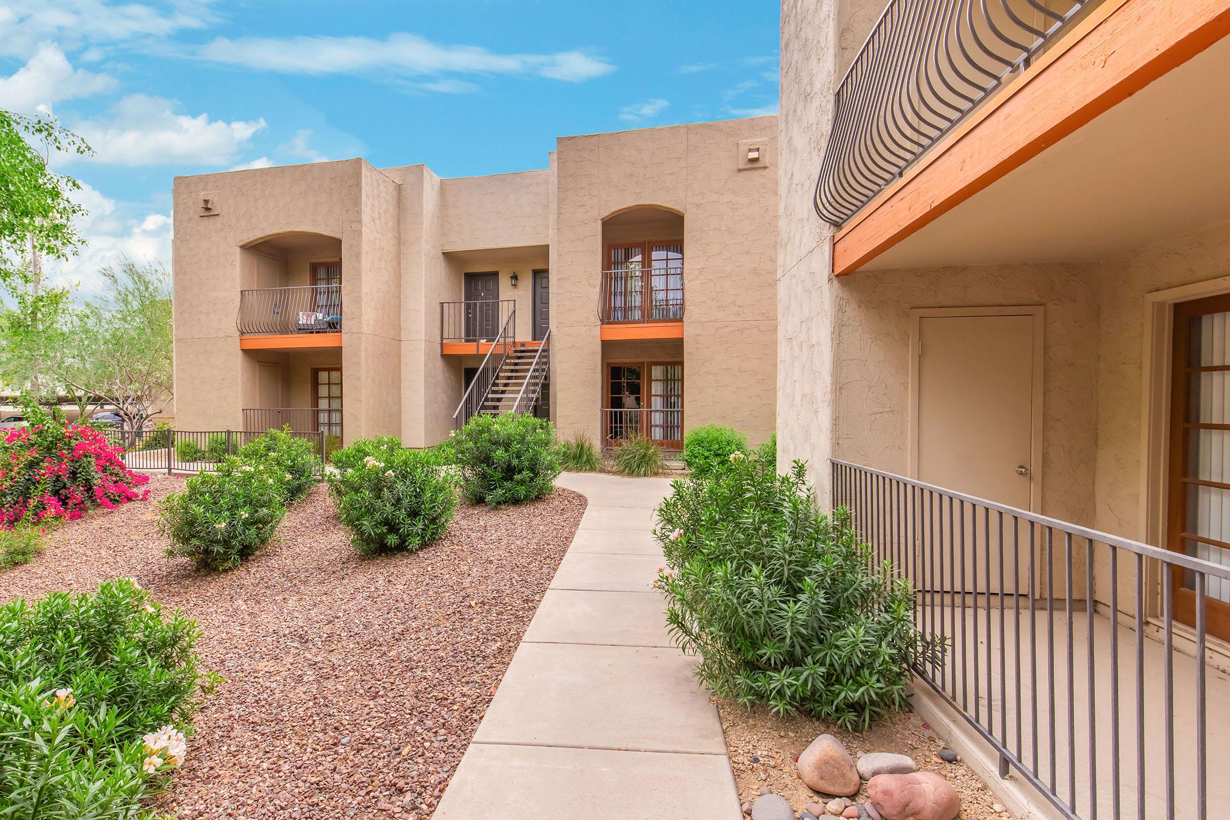 View of an apartment complex featuring beige buildings with orange accents, a landscaped pathway lined with greenery and flowers. Stairs lead to upper-level units, framed by a clear blue sky, creating a welcoming and tranquil outdoor environment.