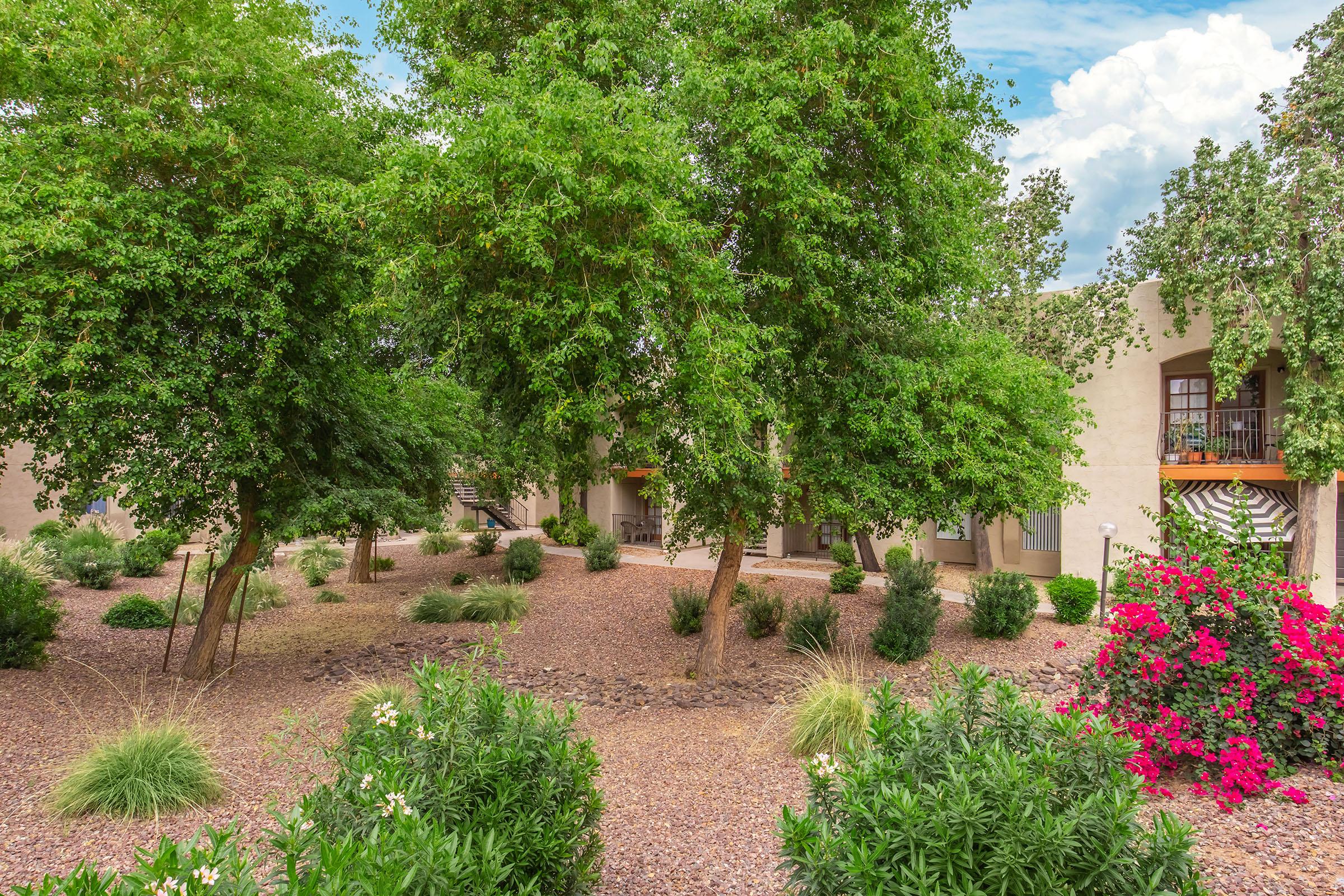 A landscaped courtyard featuring lush green trees and vibrant flower bushes. The area is adorned with gravel ground cover, creating a serene outdoor space typical of an apartment complex. In the background, residential buildings are partially visible, adding to the tranquil atmosphere.