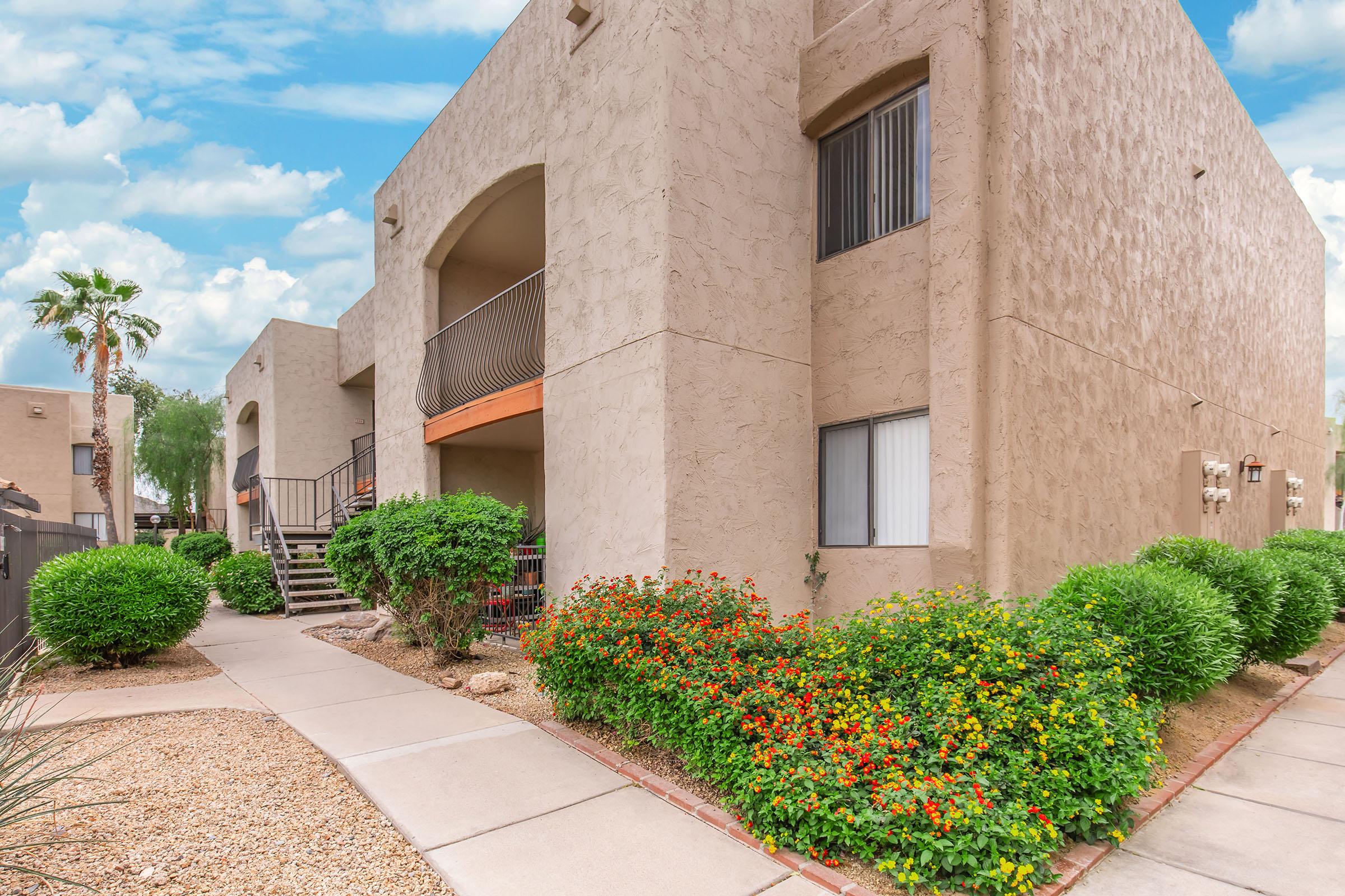 A view of a landscaped apartment complex featuring beige stucco buildings, lush green shrubs, and vibrant flower beds. The scene includes a pathway leading to the entrance, with palm trees in the background and a blue sky dotted with clouds.