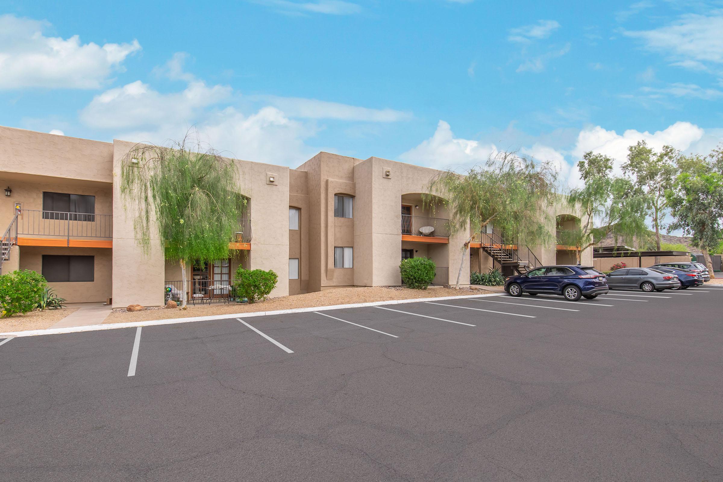 A view of a residential apartment complex featuring beige buildings with balconies, surrounded by greenery and parking spaces. A few cars are parked in front, and the sky is blue with white clouds. The area has a clean, well-maintained appearance.