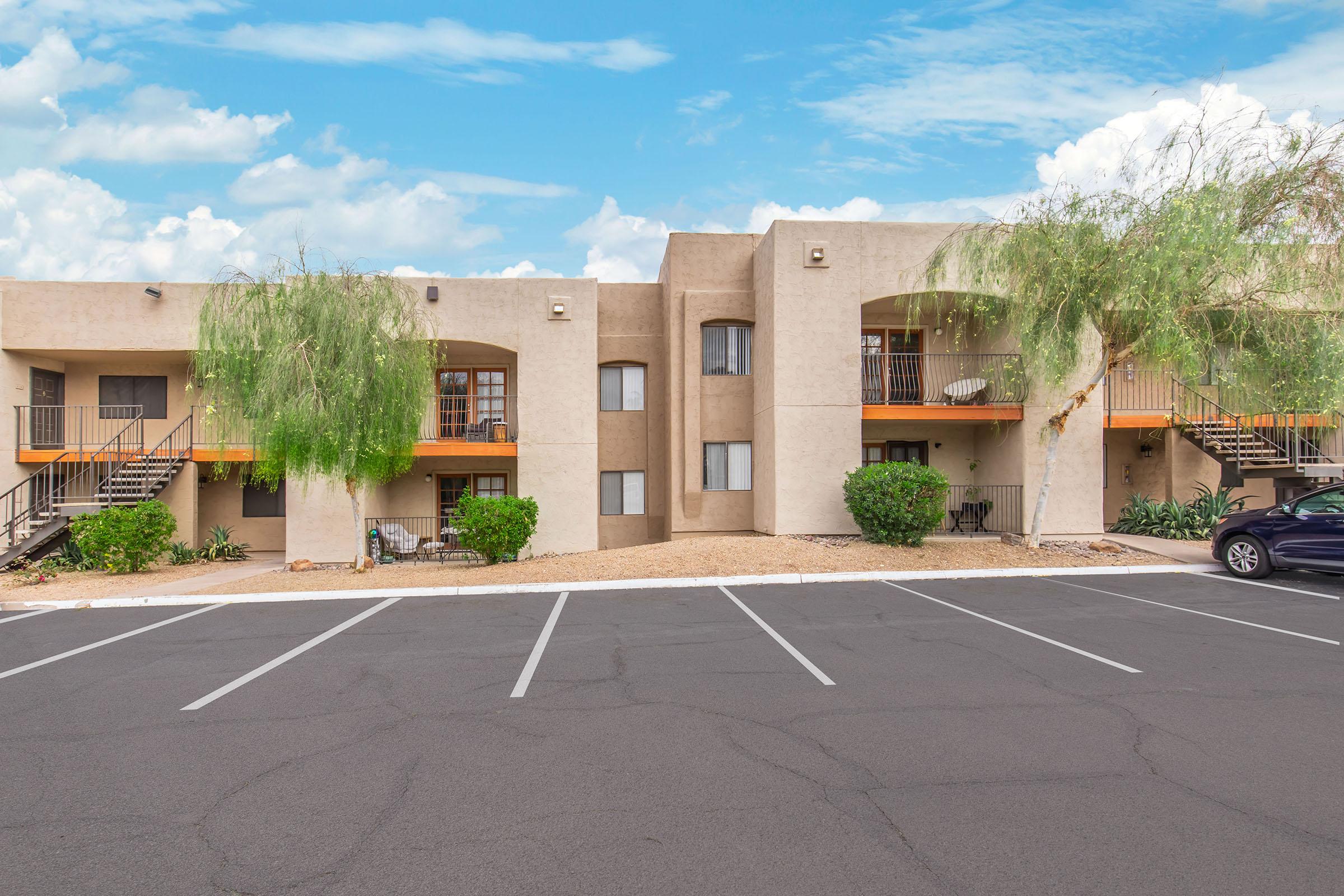A view of a multi-unit apartment complex featuring beige exterior walls, multiple balconies, and landscaped greenery. The parking lot in front has several marked spaces, and the sky is partly cloudy.