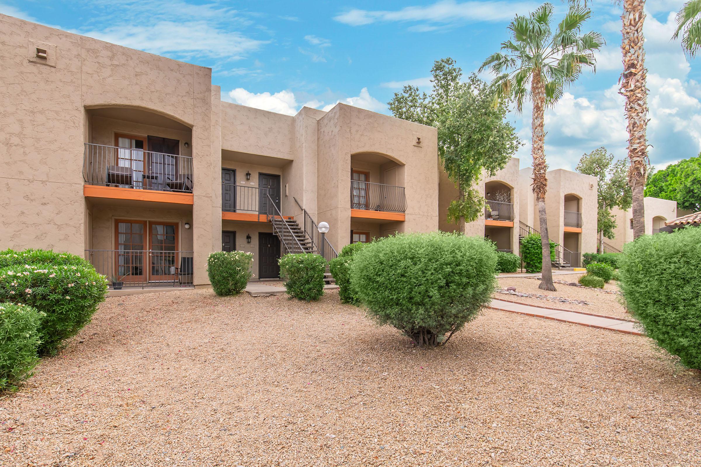 Exterior view of a multi-unit apartment complex featuring beige stucco buildings with balconies. Lush shrubs and palm trees surround the buildings, and a gravel pathway leads to the entrance. The sky is clear with scattered clouds, creating a pleasant outdoor setting.