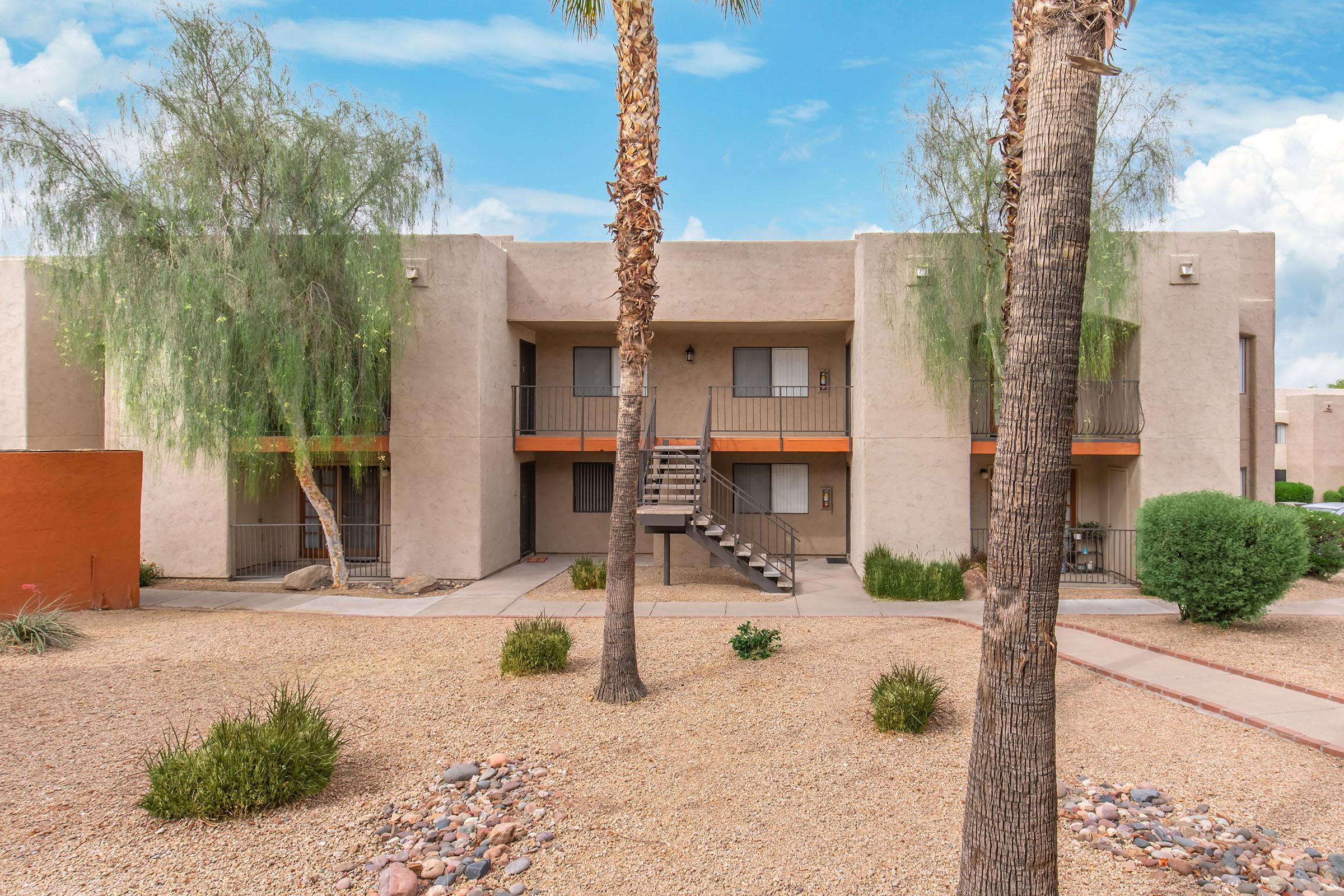A two-story apartment building with a textured exterior in a desert landscape. It features two entrances with staircases, surrounded by palm trees and shrubs. The ground is covered with gravel and small rocks, creating a low-maintenance outdoor area. The sky is bright with scattered clouds.