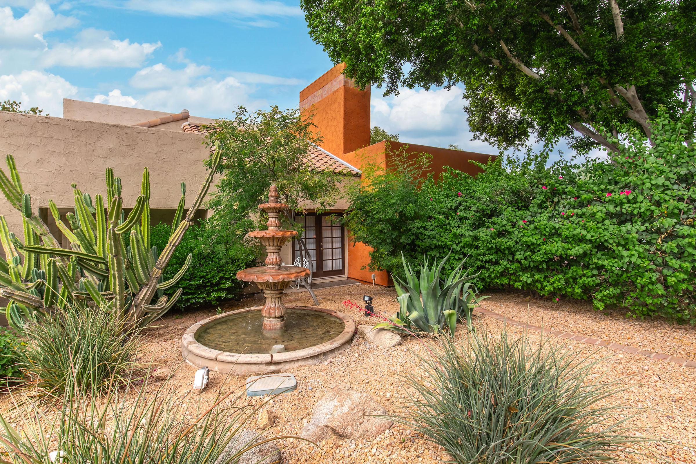 A desert garden with a central tiered fountain surrounded by various cacti and lush greenery. The building has a warm orange wall, with a tiled roof peeking through the plants. The scene is bright and inviting, featuring a clear blue sky in the background.