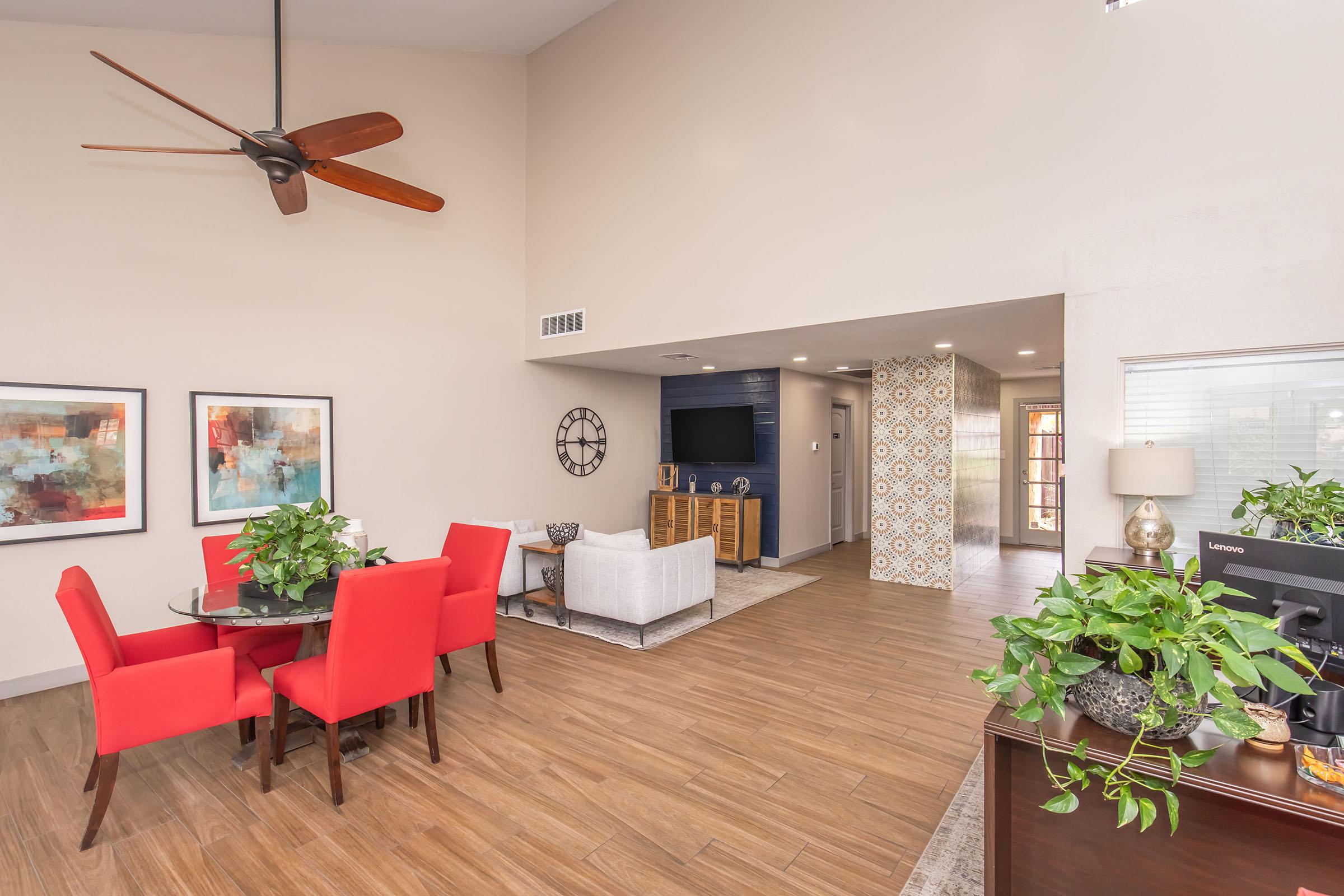 A spacious interior living area featuring a light-colored wall, wooden flooring, and modern decor. There's a round dining table with red chairs, a cozy sitting area with a white couch, and a TV mounted on the wall. Decorative plants are placed throughout the space, adding greenery to the design.