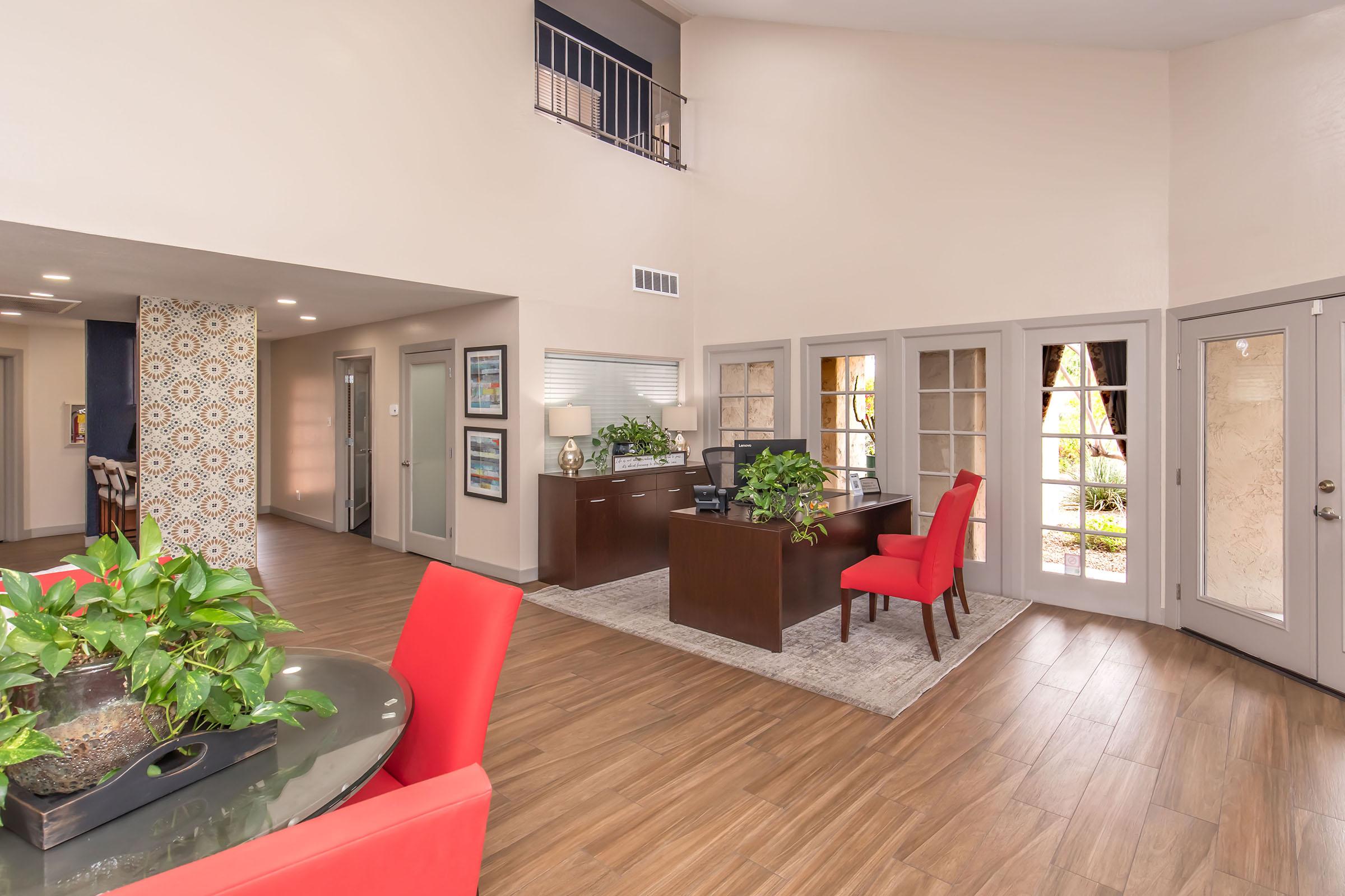 Modern reception area featuring a dark wood desk with a computer and greenery, surrounded by light-colored walls. There are two red chairs and a circular dining table with plants. Glass doors lead to an outdoor space, and patterned wallpaper adds visual interest to the design.