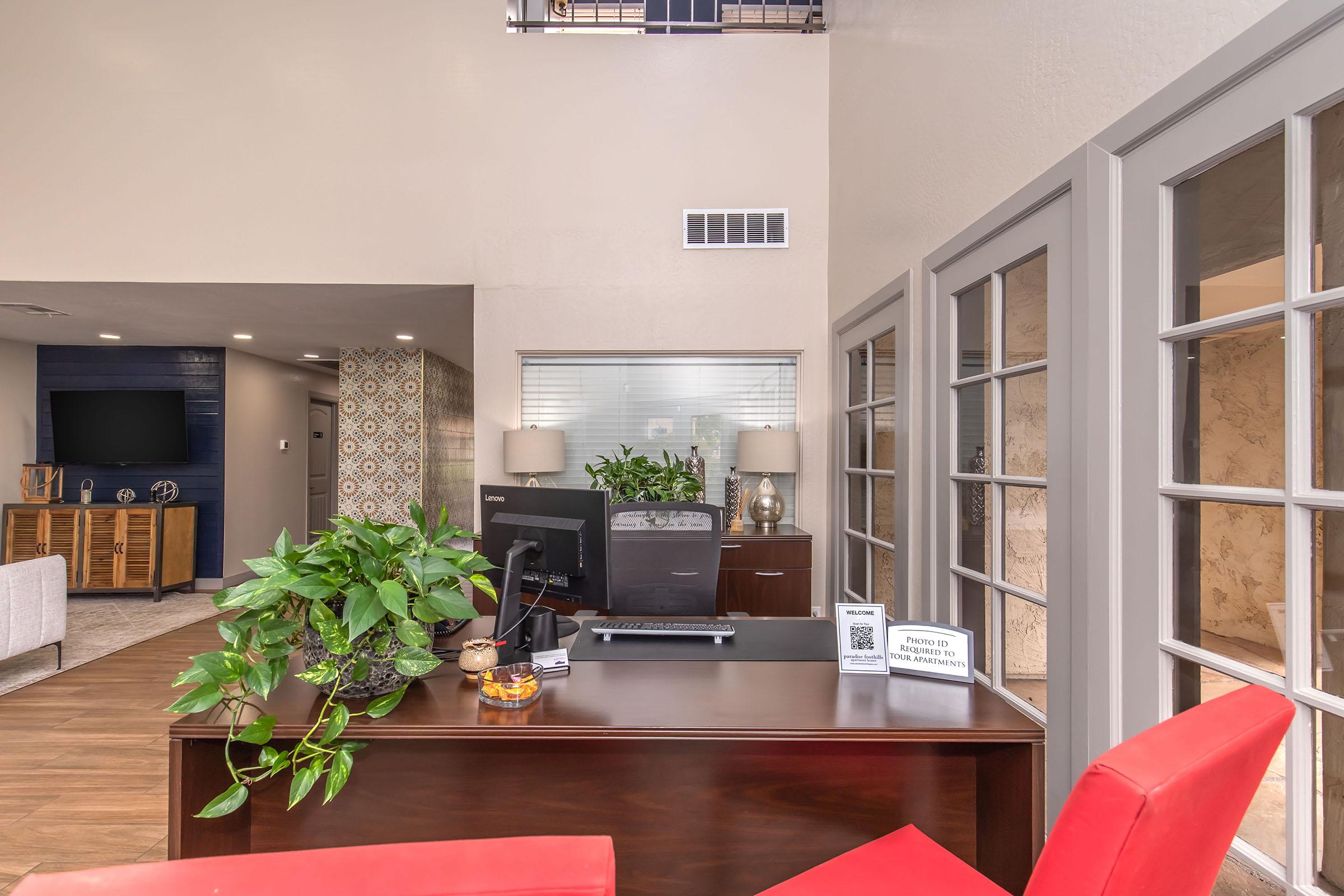 A modern office reception area with a wooden desk featuring a computer and small decorative items. Potted plants are arranged on the desk, and there's a welcoming ambiance with neutral-colored walls. A seating area with light-colored furniture is visible in the background, along with a TV and patterned wall design.