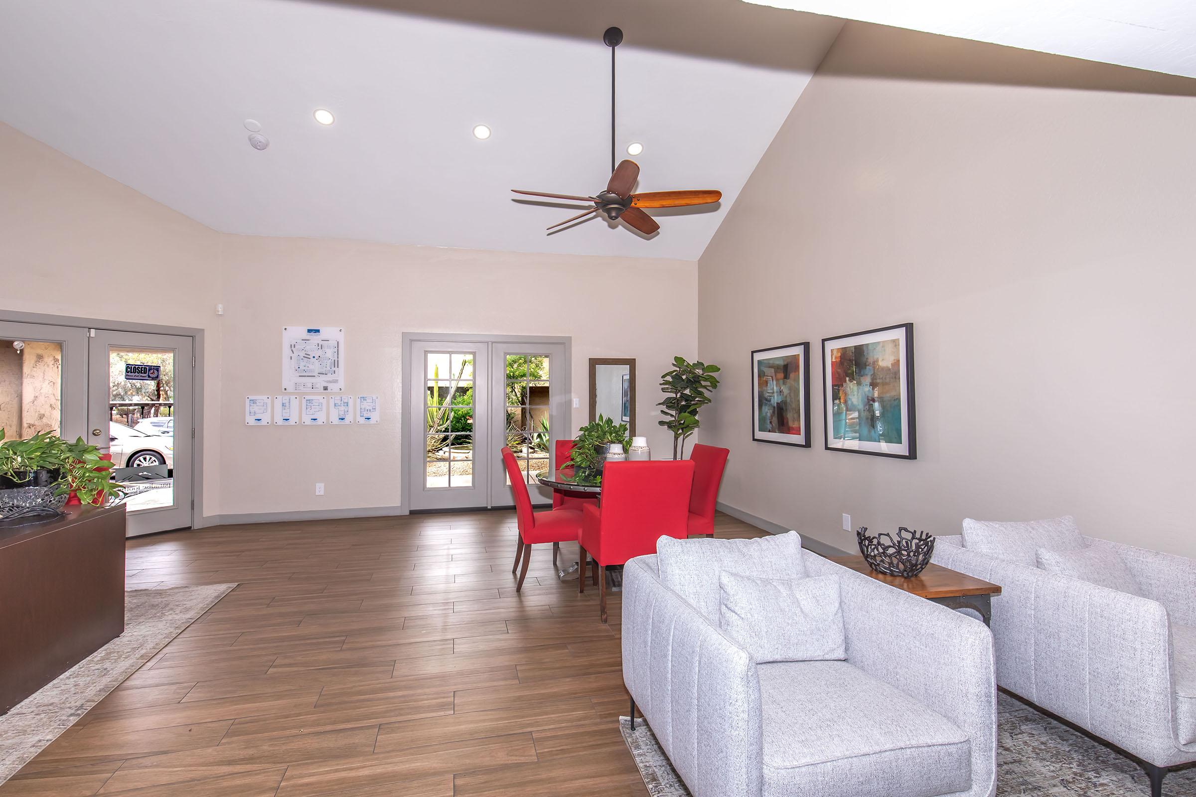 Interior of a modern reception area featuring two gray armchairs, a wooden reception desk, and a dining set with red chairs. Large windows allow natural light in, and decorative plants add a touch of greenery. The ceiling is high with a ceiling fan, creating an inviting atmosphere.