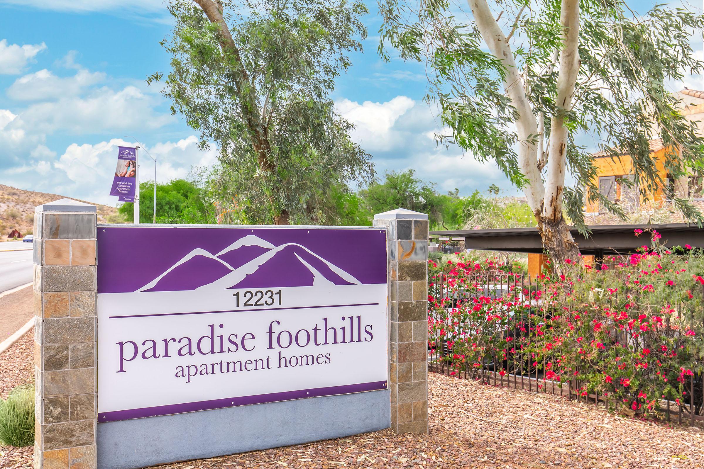 Sign for Paradise Foothills Apartment Homes, featuring the address "12231." The sign is prominently displayed with purple and white colors, set against a backdrop of flowering plants and trees, under a partly cloudy blue sky.