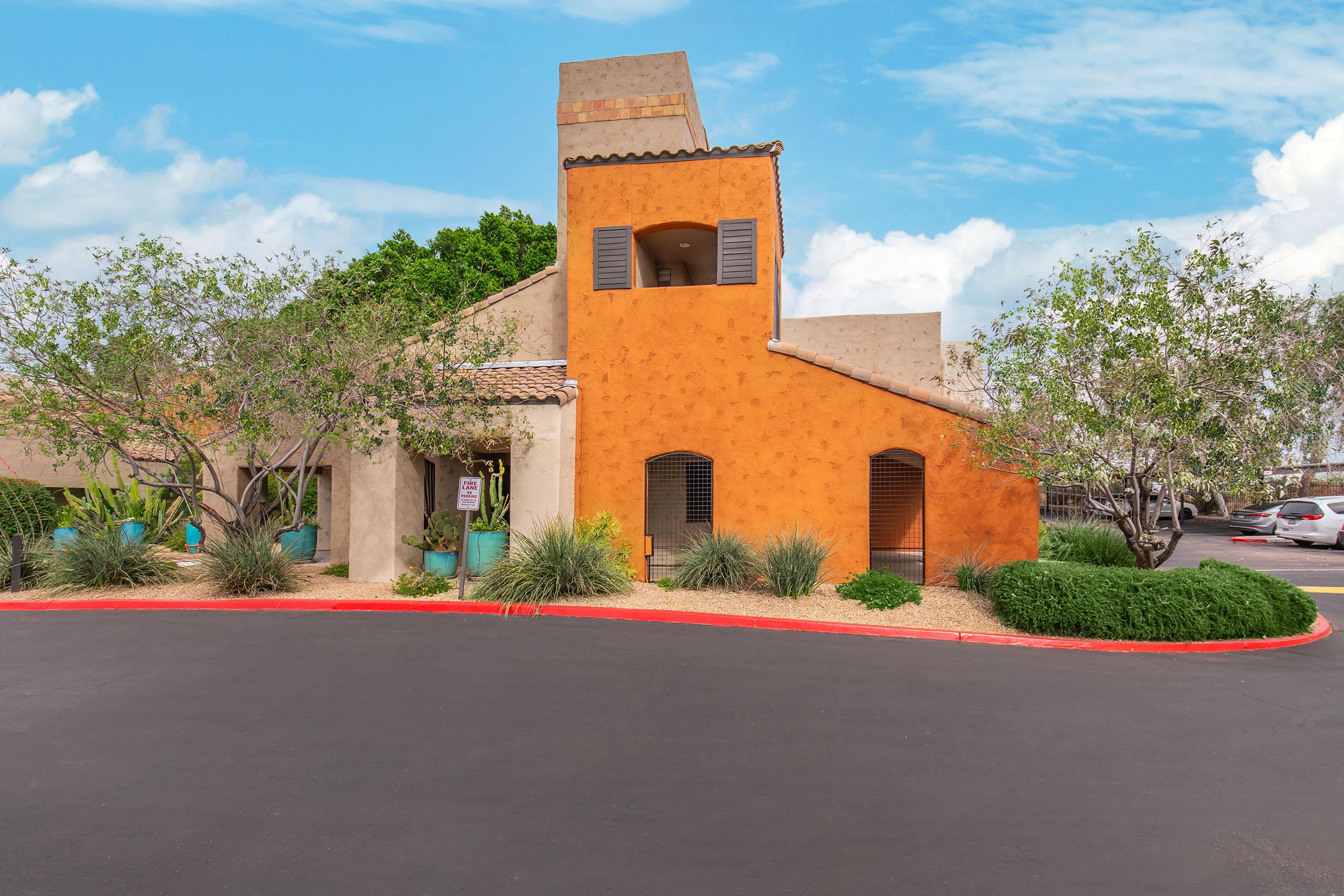 Vibrant adobe-style building with orange and tan walls, featuring a unique tower and blue shutters. Surrounding landscape includes small trees and shrubs, and a curved driveway with a smooth asphalt surface. The sky is bright with scattered clouds, creating a welcoming atmosphere.