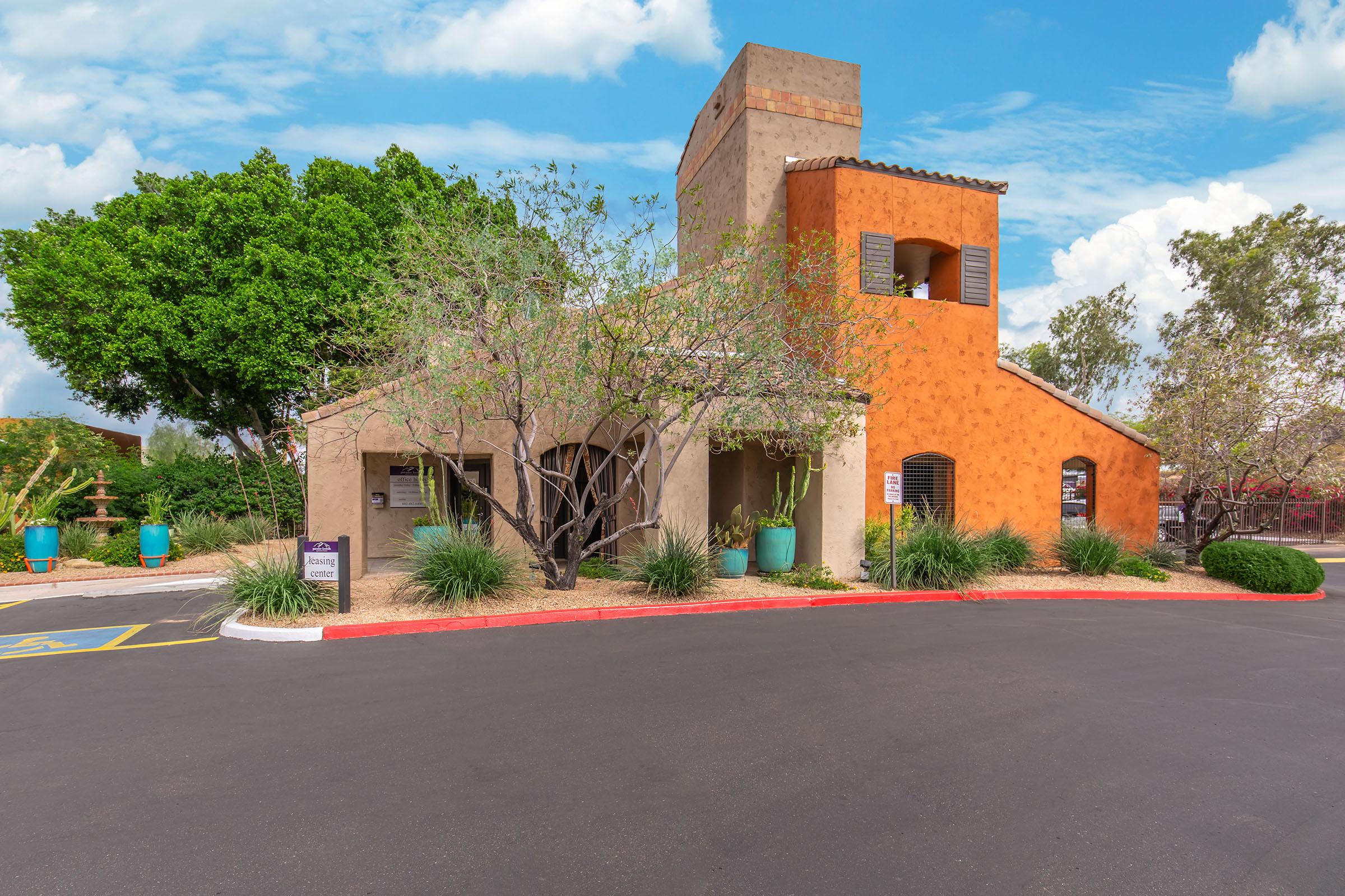 A modern adobe-style building with a warm orange exterior, surrounded by lush greenery and desert landscaping. The structure features large windows, a bell tower, and is set against a blue sky with scattered clouds. There are marked parking spots and decorative planters visible in the foreground.