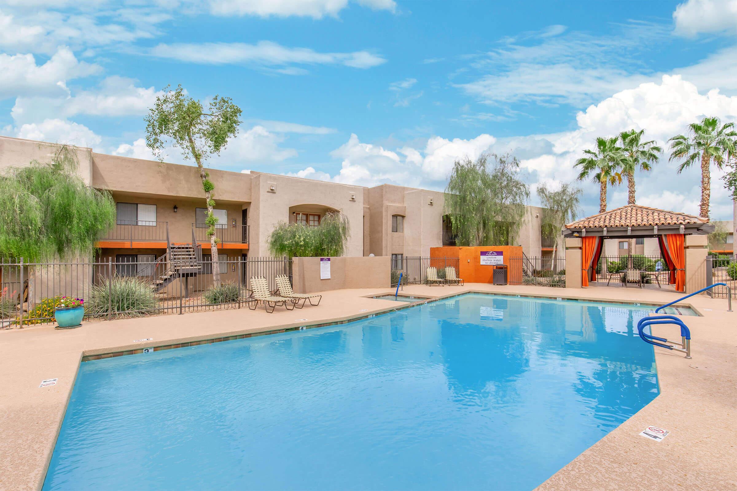 A sunny outdoor pool area with a clear blue pool surrounded by lounge chairs. In the background, there are apartment buildings and palm trees. A cabana with orange drapes adds a cozy touch to the space. The sky is partly cloudy, creating a pleasant atmosphere.
