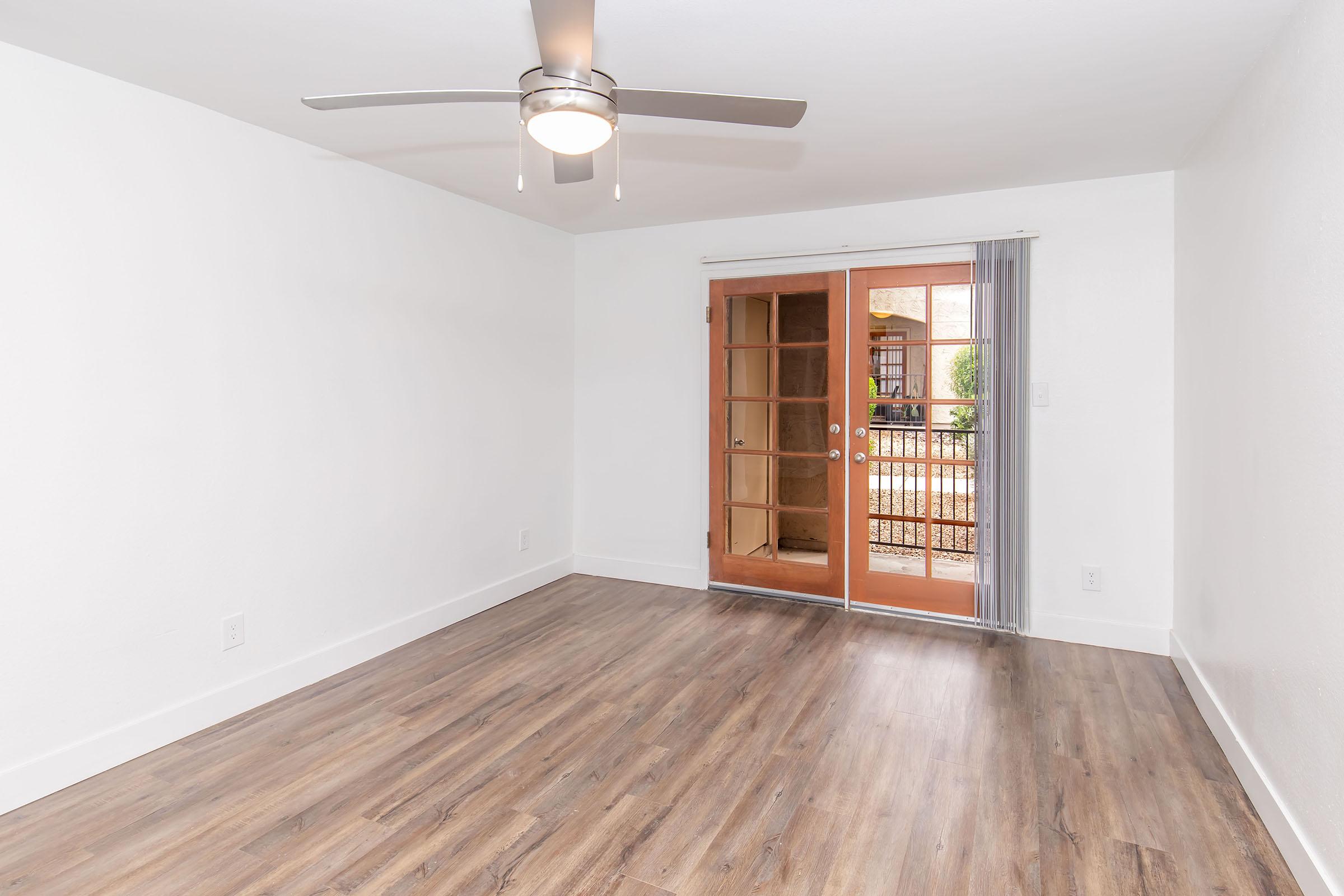 A spacious, empty room with light wood flooring, a ceiling fan, and large sliding glass doors that open to a patio or outdoor area. The walls are painted white, creating a bright and airy atmosphere. Natural light floods in through the doors, enhancing the room's openness.