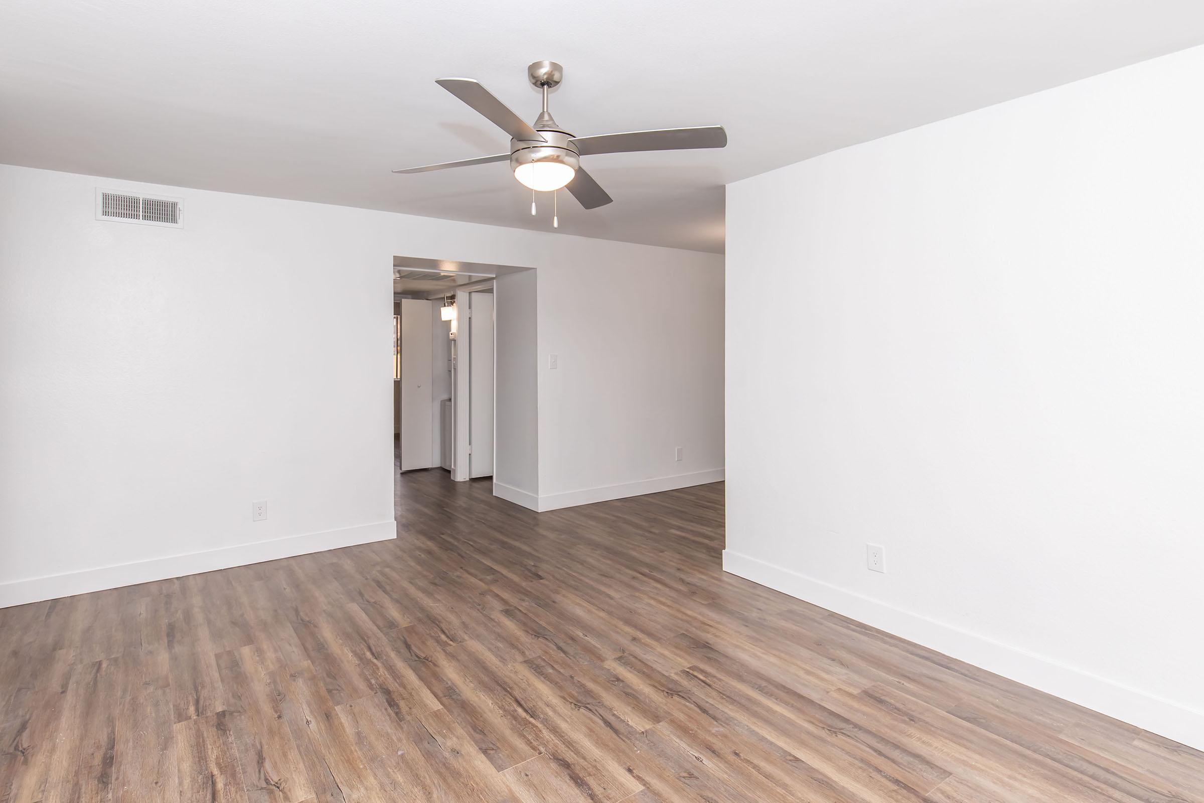 A spacious, modern living area featuring light-colored walls and hardwood-style flooring, with a ceiling fan providing light and ventilation. A doorway leads to an adjacent room, suggesting an open-plan layout. The space is unfurnished, ideal for customization.