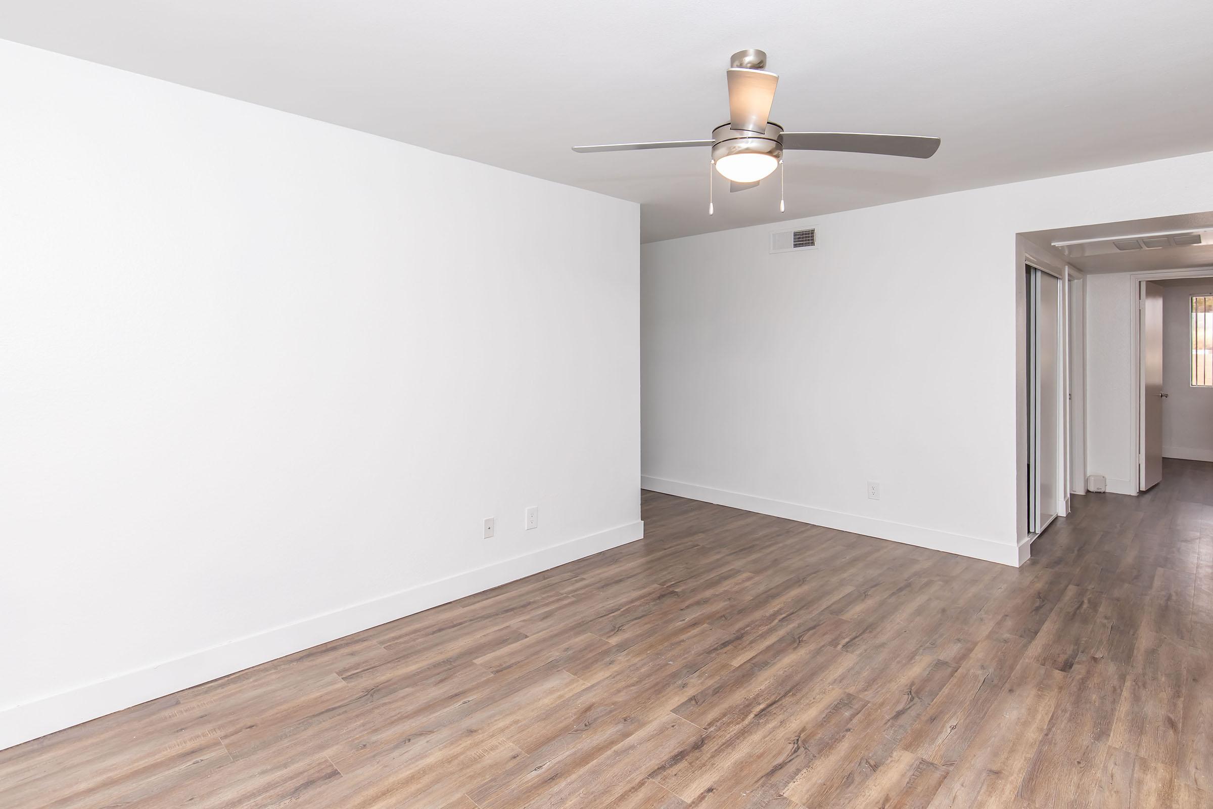 A spacious, empty room with light-colored walls and a wooden floor. There is a ceiling fan with light fixtures, and an opening to a hallway on the right side. The lighting is bright, creating a clean and airy atmosphere, suitable for various interior designs and furnishings.